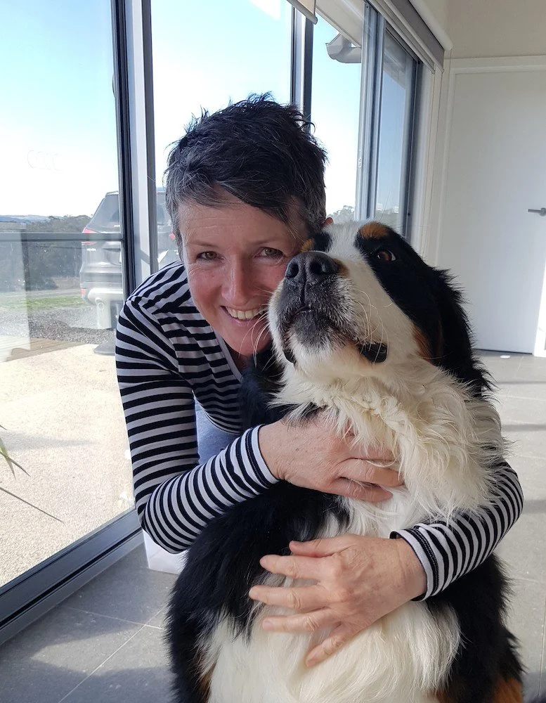 A woman in a black and white striped shirt hugging a large Bernese Mountain Dog inside a house near glass sliding doors with a view outside.