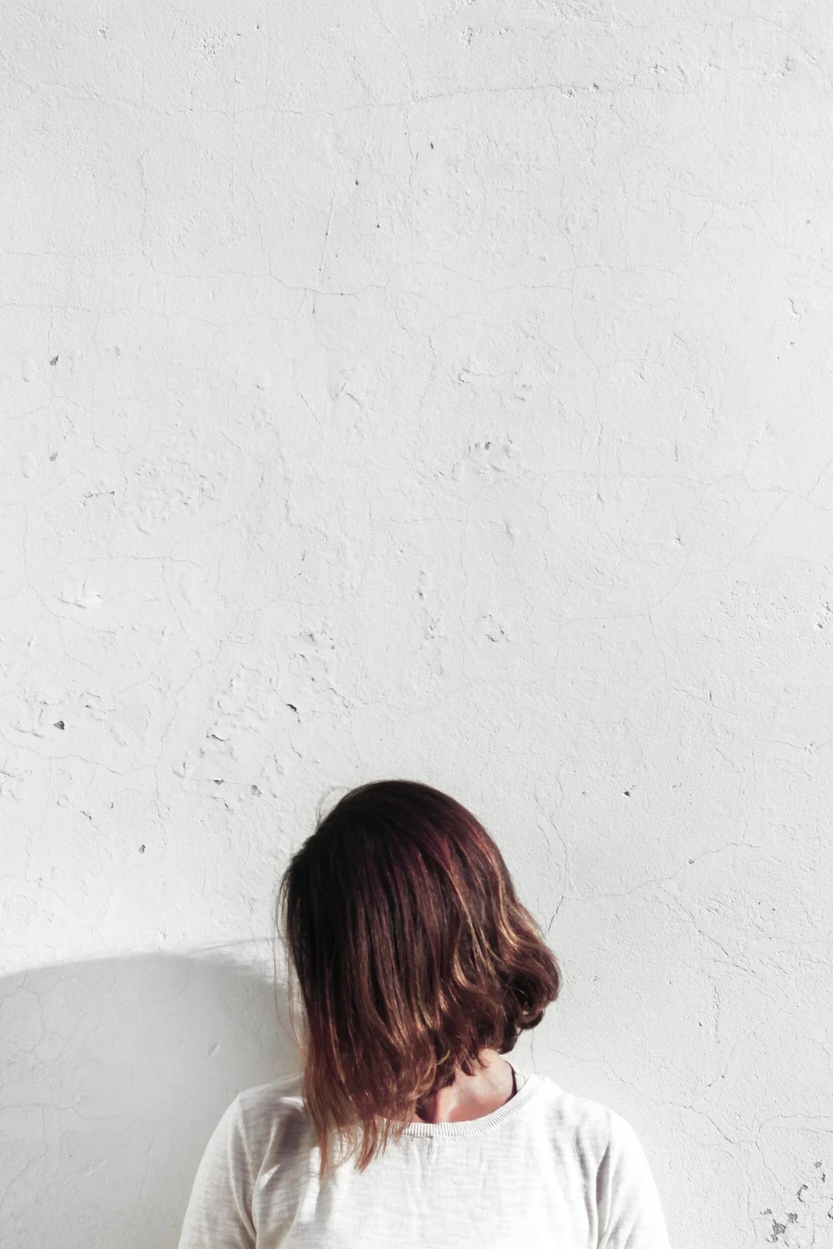 A person with shoulder-length brown hair with highlights, facing away, wearing a light-colored sweater, standing against a textured white wall.