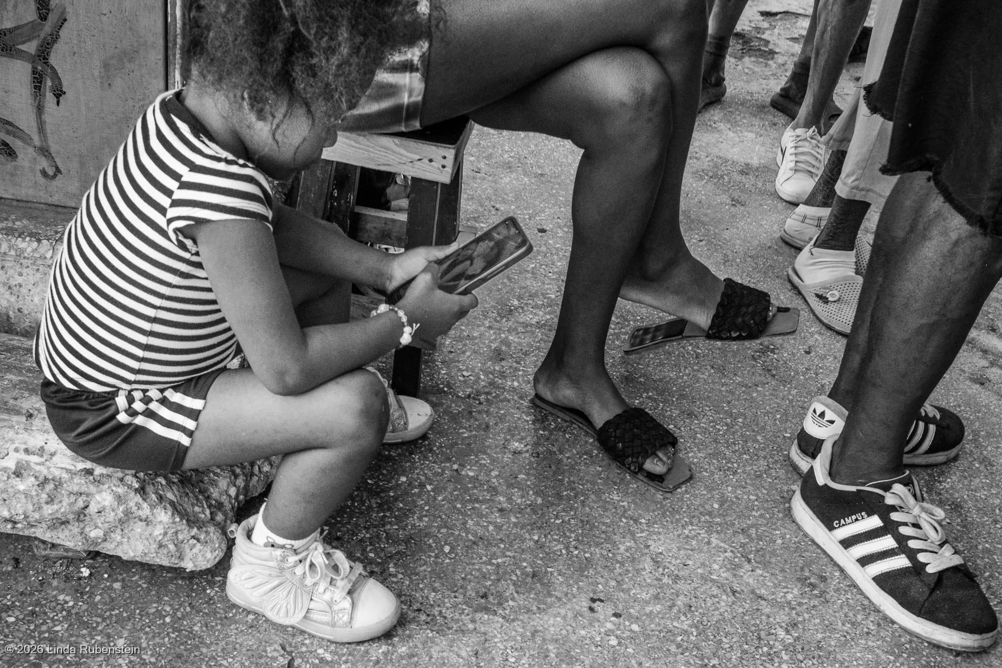 Young girl on cell phone surrounded by adult legs and feet Havana Cuba