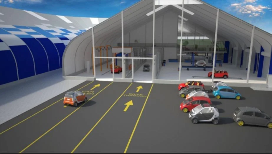 A parking lot with designated spaces and arrows leading into a large vehicle maintenance or service garage with a glass front. Several cars are parked outside and inside the garage, with a few visible in the background.