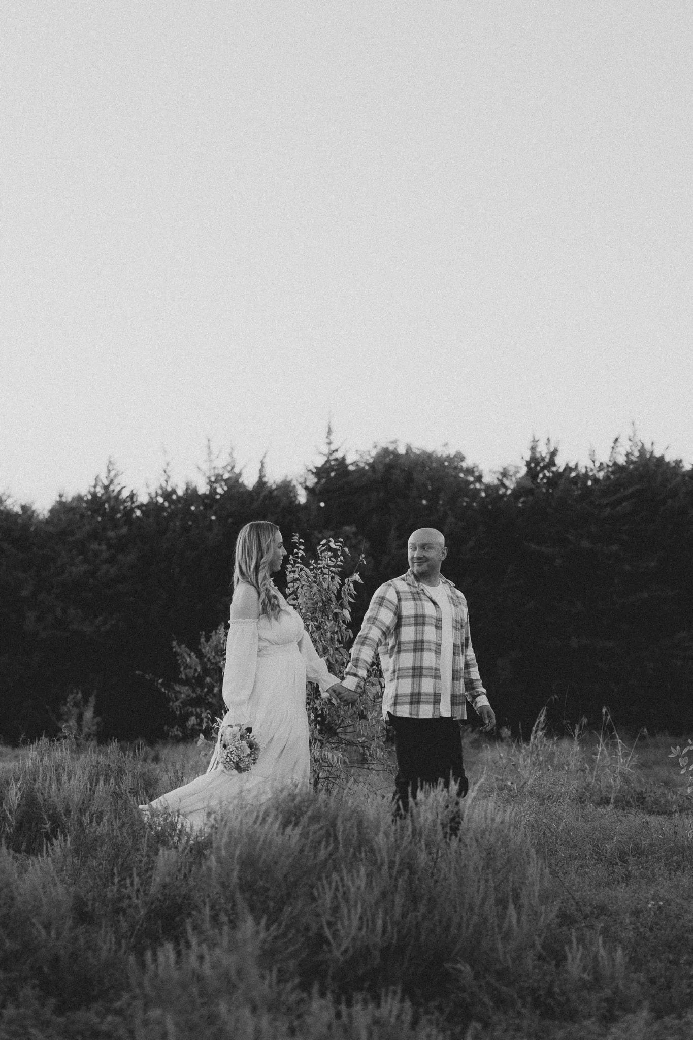 A black-and-white photo of a couple holding hands in a field, with trees in the background.