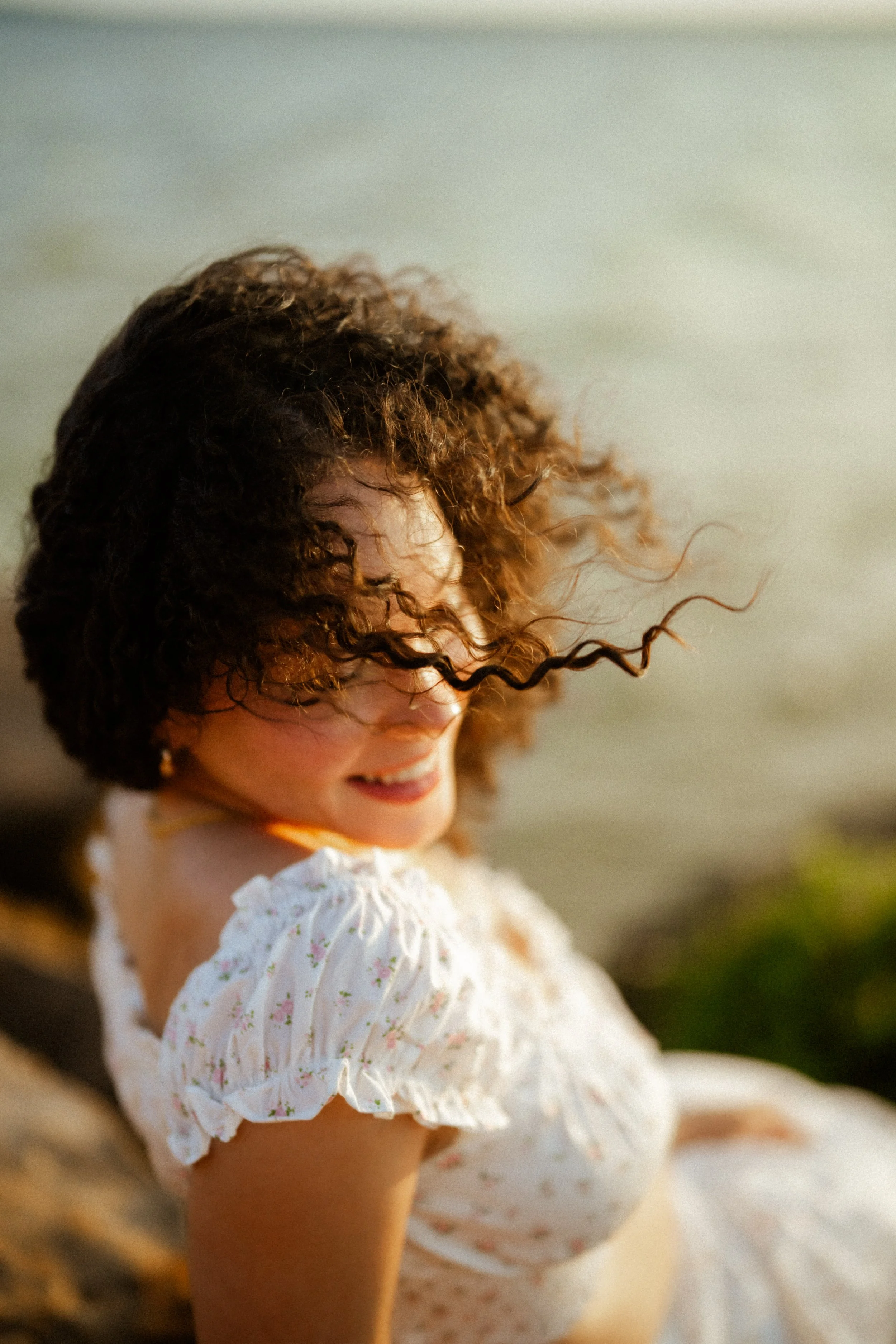 A woman with curly brown hair smiling and looking down, wearing a white floral dress, outdoors near water.