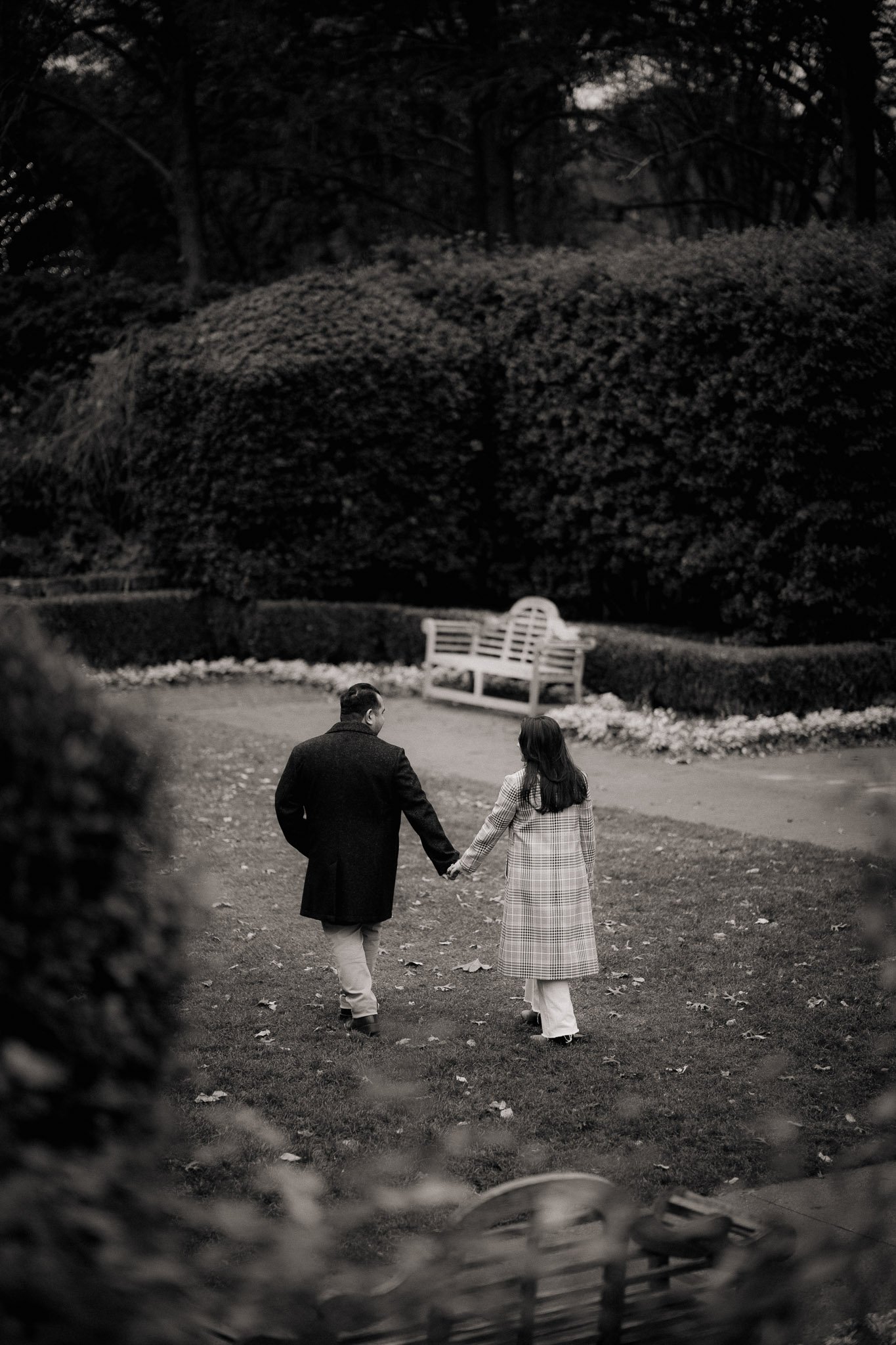 A black and white photo of a couple holding hands and walking away on a park pathway surrounded by trees and bushes, with a bench in the background.