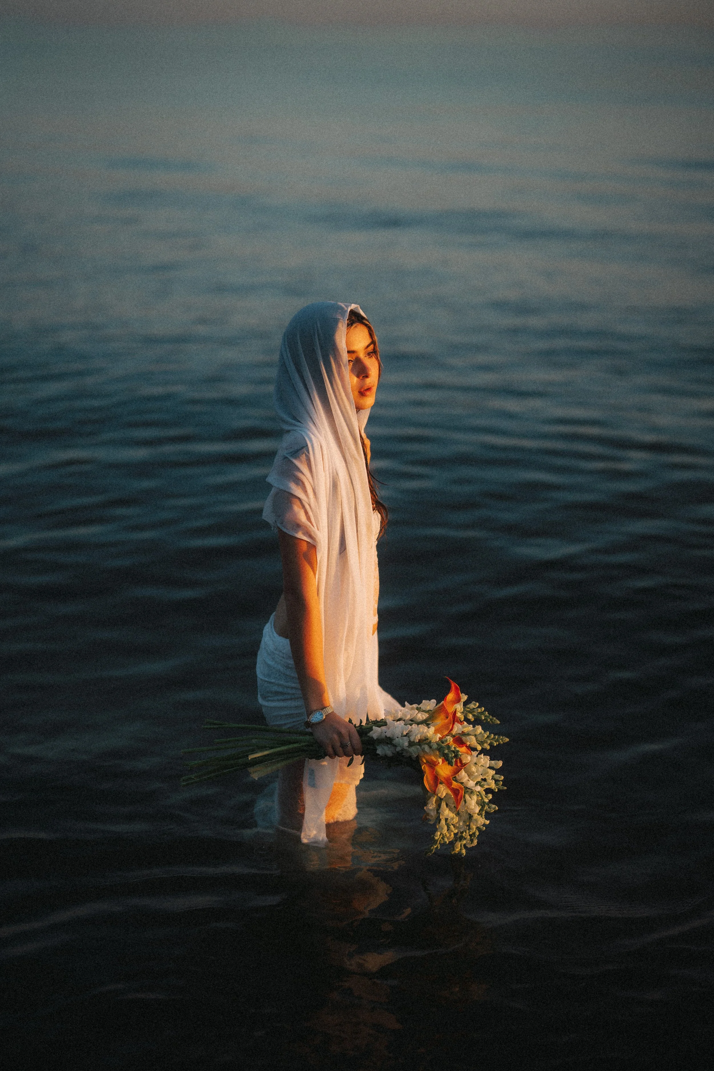 A woman standing waist-deep in water, wearing a white dress and headscarf, holding a bouquet of flowers during sunset.