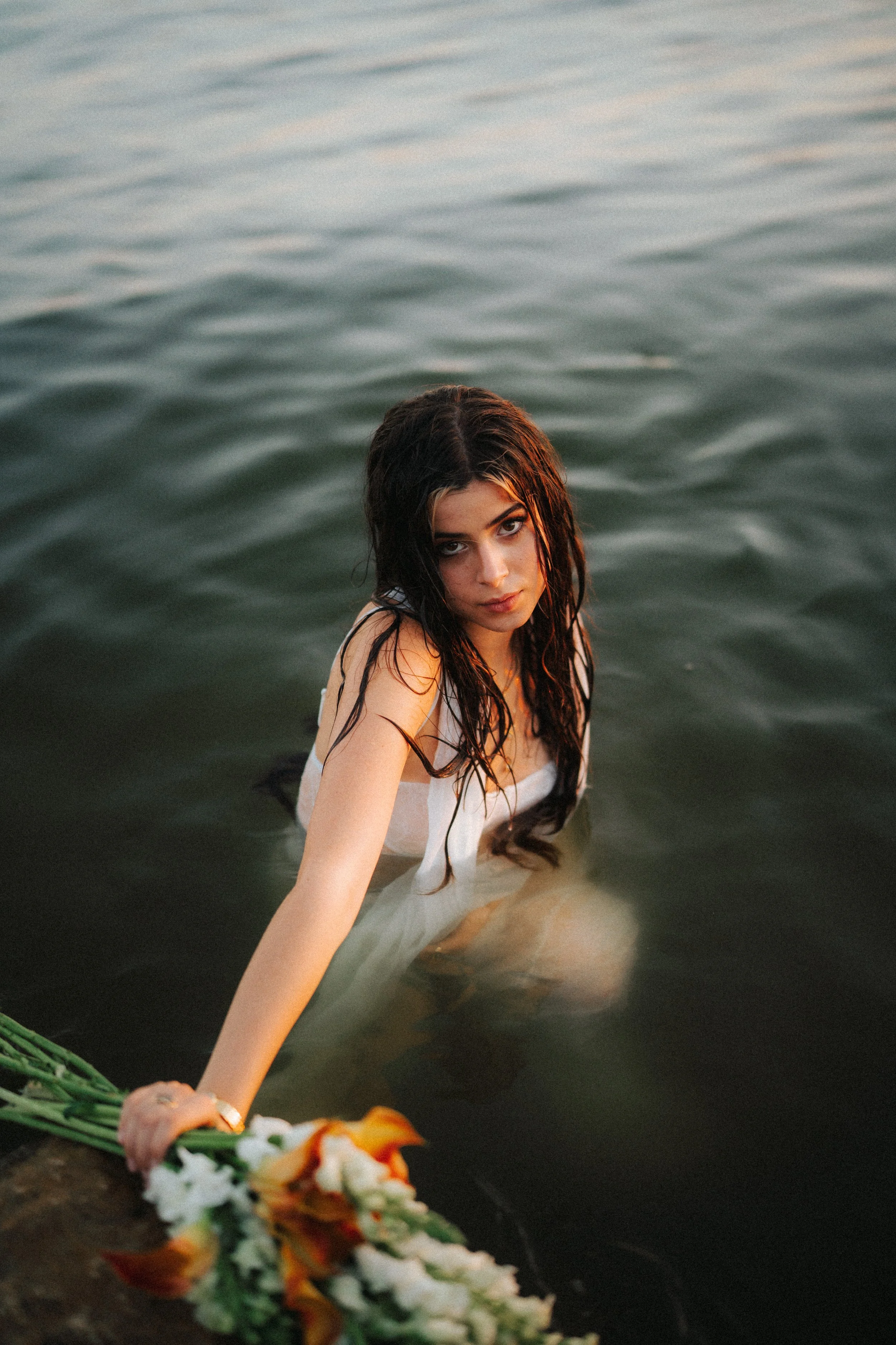 Young woman with wet dark hair sitting in water, holding a bouquet of flowers, looking at the camera.