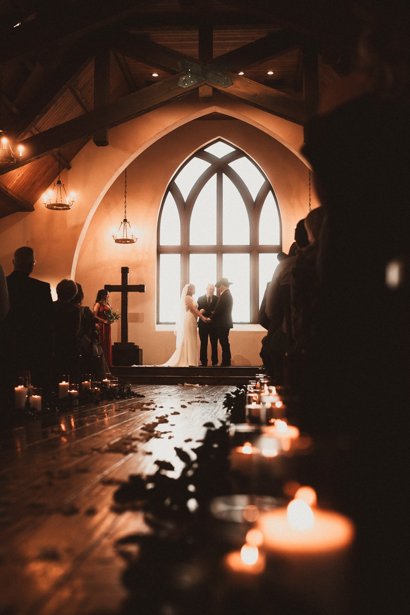 A bride and groom holding hands at the altar of a church during their wedding ceremony, with a priest in the background, city fog visible through large arched windows, candles along the aisle, and guests seated on either side.