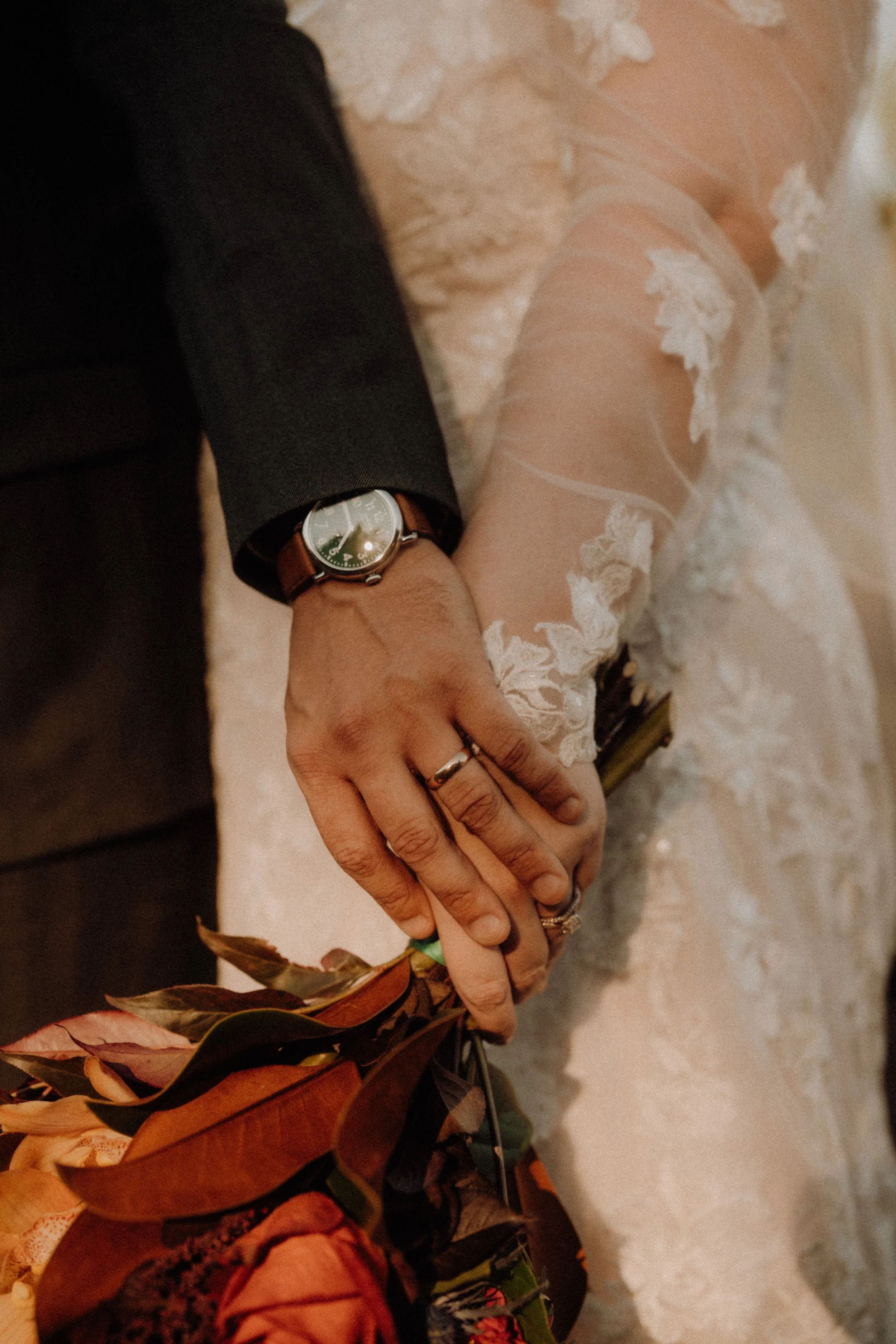 Close-up of a newlywed couple's hands intertwined, showing rings, with a bouquet of flowers, on the bride's lace dress and the groom's dark suit.