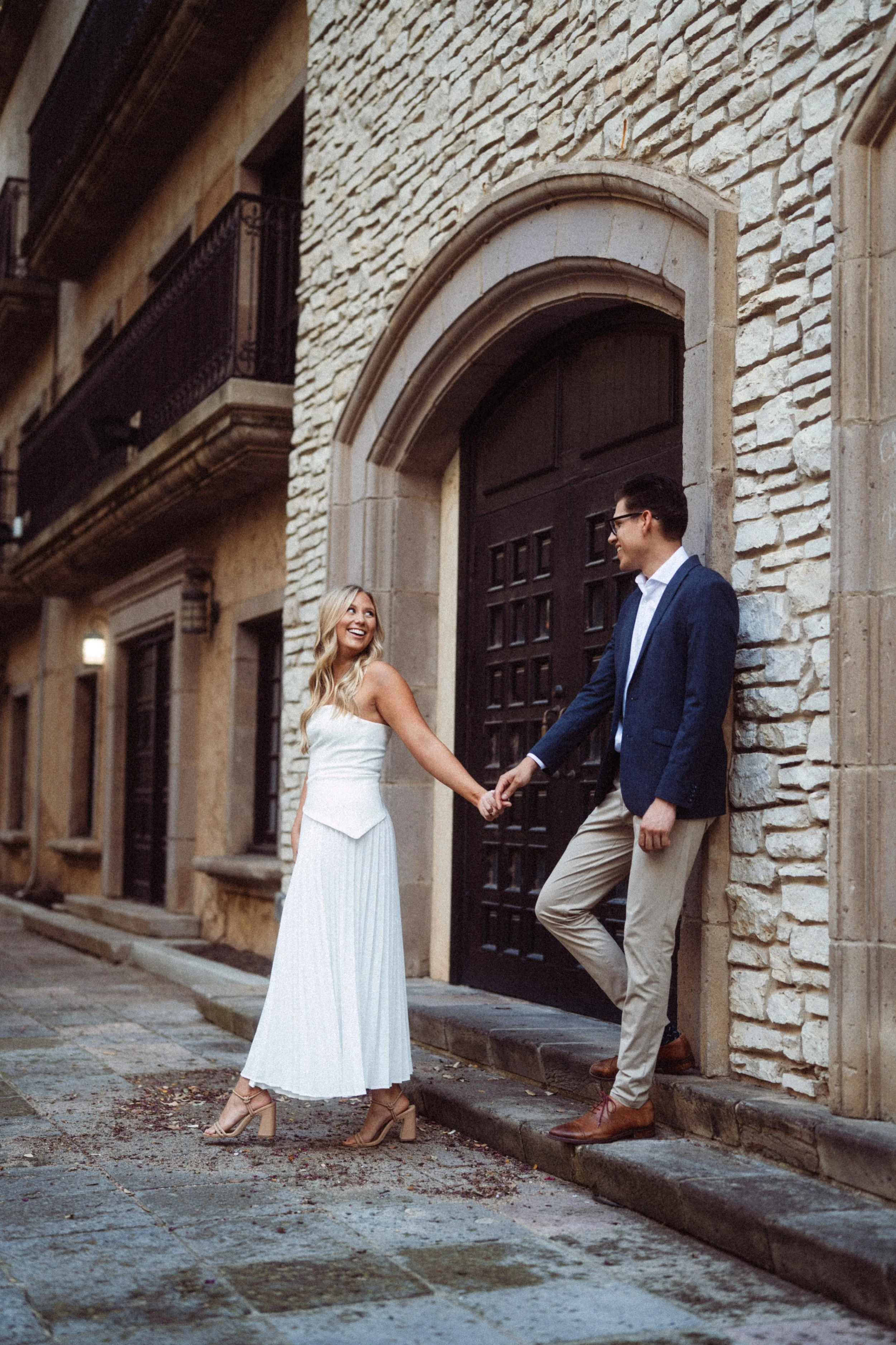  A joyful, candid engagement photograph of Alaina and Andres holding hands and smiling at each other in front of a rustic wooden door and stone wall. This romantic portrait was captured during their engagement session at the European-inspired Mandala