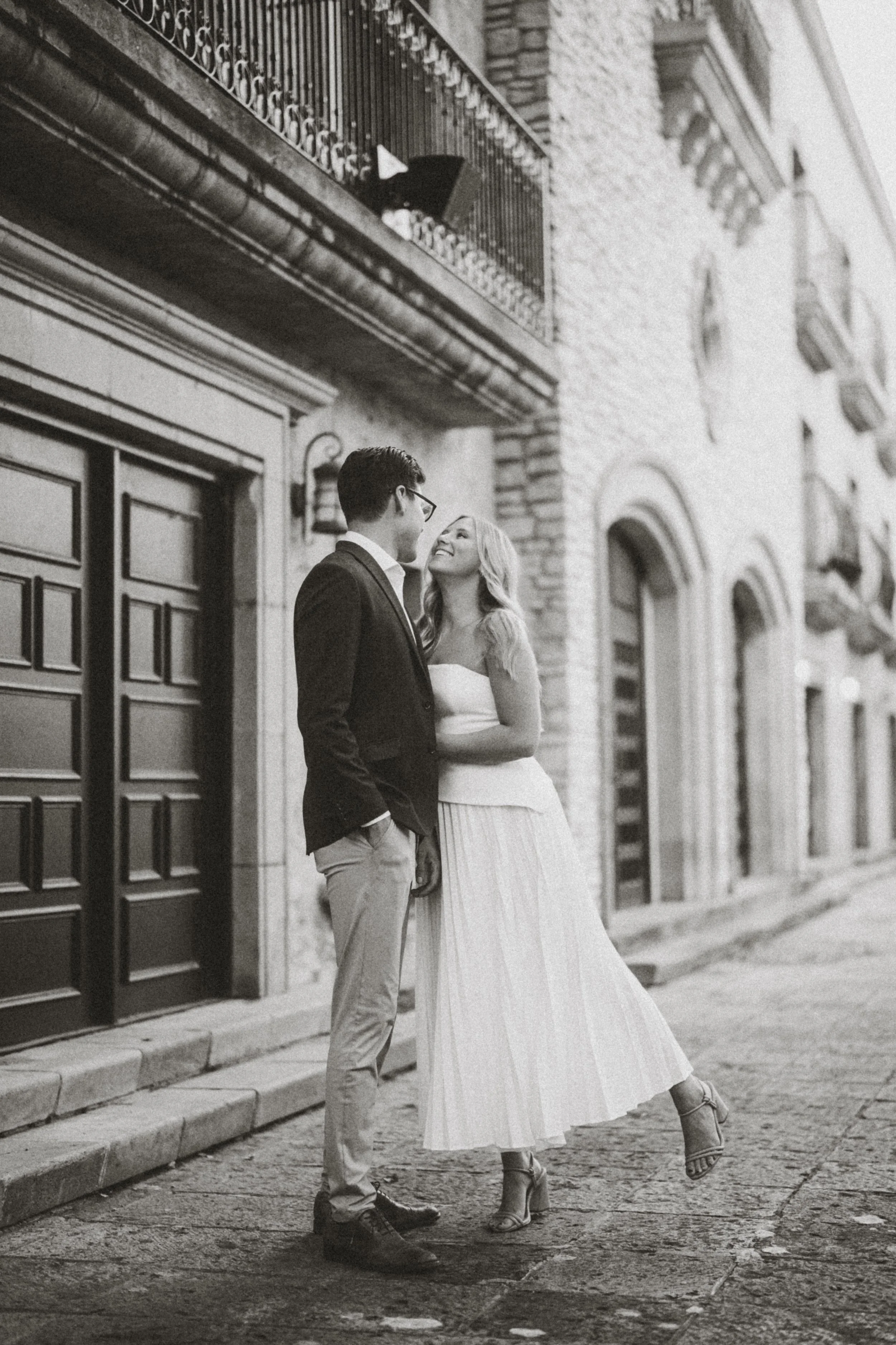  A timeless black and white engagement photograph of Alaina and Andres standing together and smiling on a cobblestone path. This romantic portrait was captured during their session at the European-style Mandalay Canal in Irving, Texas. The image show