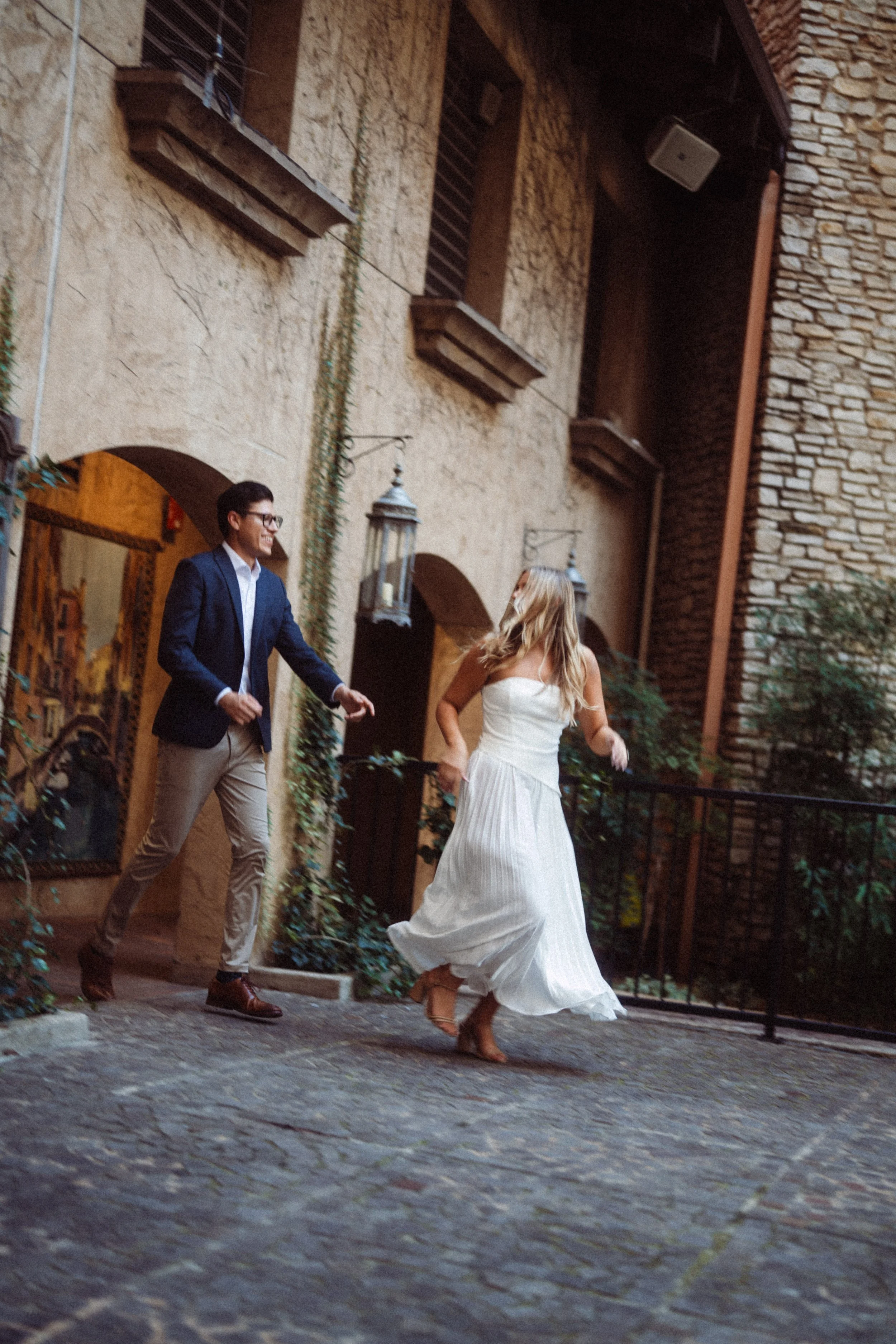  A candid, joyful engagement photograph of Alaina twirling in her white dress while Andres walks towards her with a smile. This playful moment was captured on the cobblestone walkways of the Mandalay Canal in Irving, Texas, against a backdrop of Old 