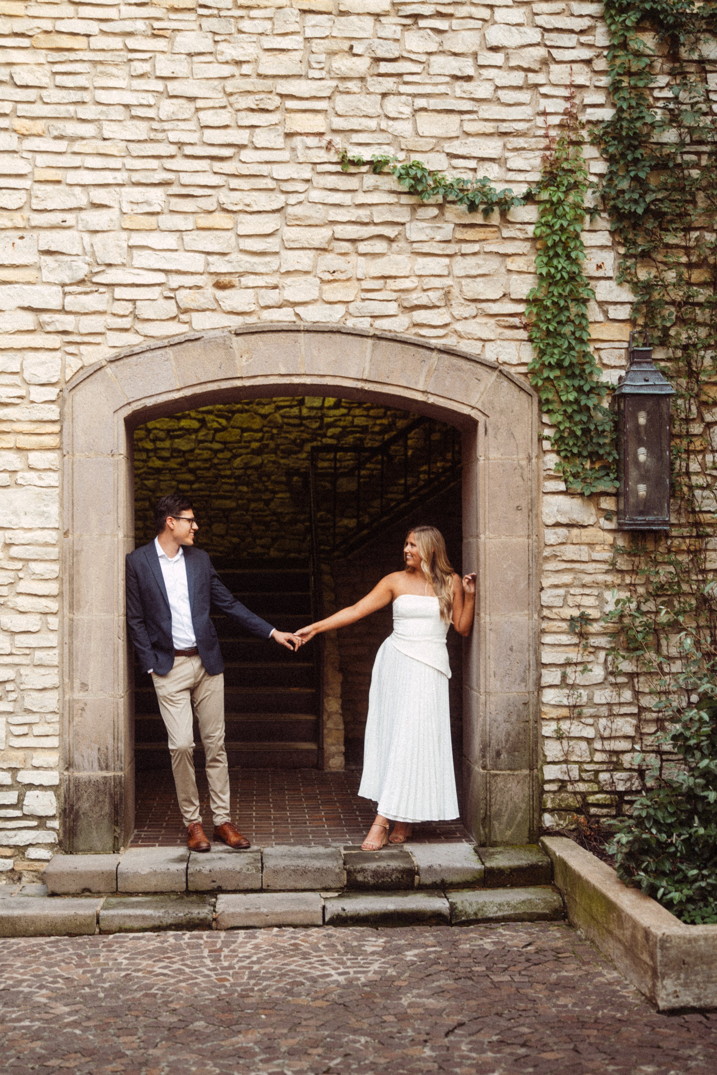  A romantic engagement photograph of Alaina and Andres holding hands and smiling at each other under a rustic stone archway covered in ivy. This timeless portrait was captured during their session at the European-style Mandalay Canal in Irving, Texas
