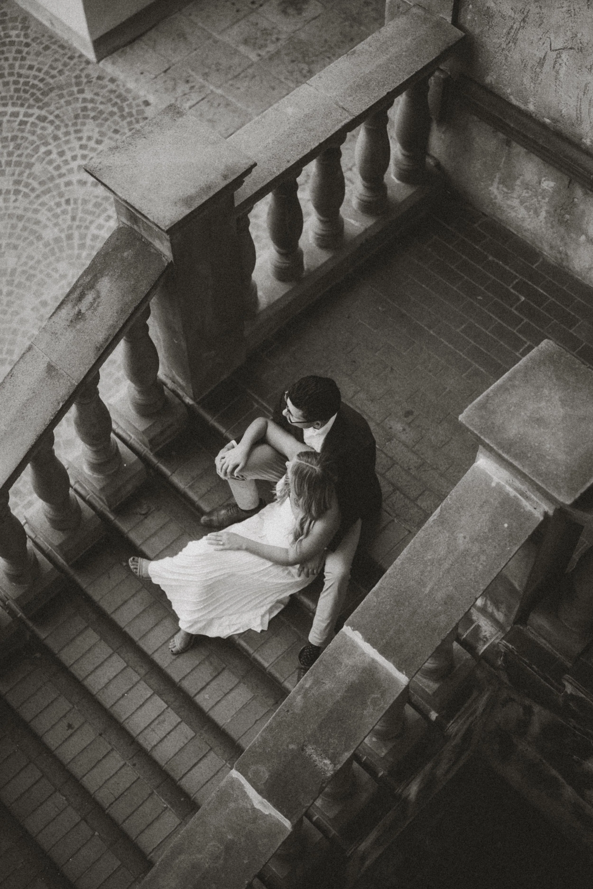  A timeless black and white overhead photograph of Alaina and Andres sitting together on a grand stone staircase, captured from a high angle. This artistic and intimate engagement portrait was taken at the Mandalay Canal in Irving, Texas, and feature