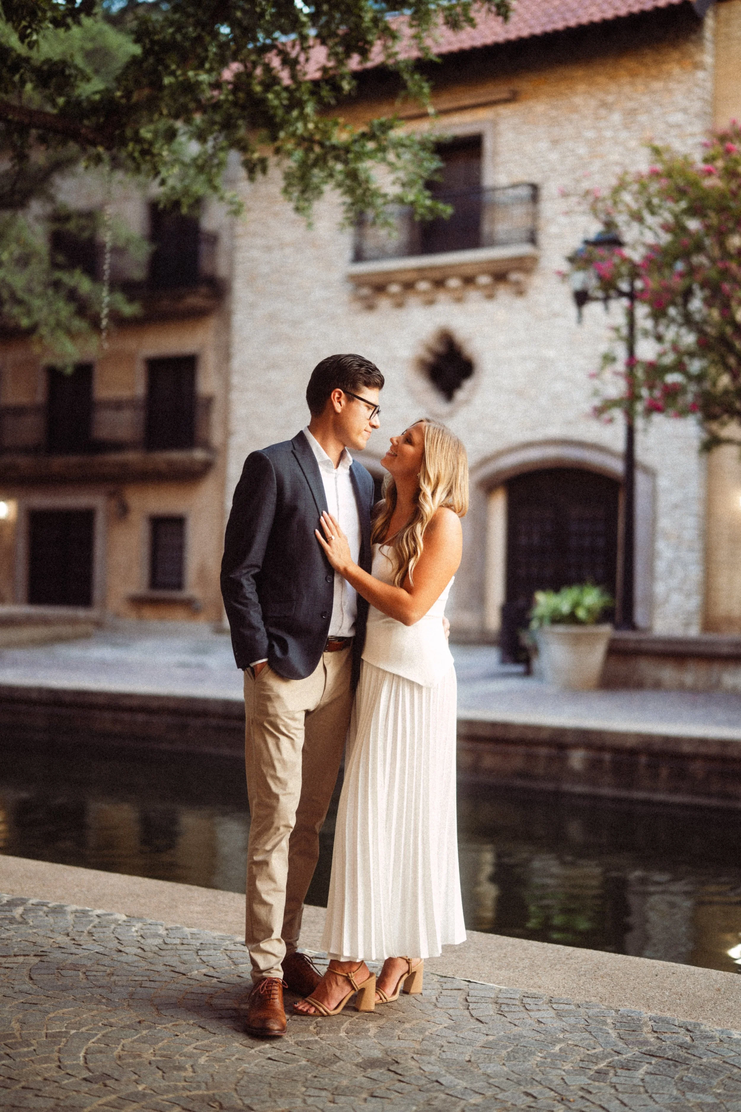  A romantic engagement photograph of Alaina and Andres standing by the water's edge, looking lovingly into each other's eyes. The session took place at the Mandalay Canal in Irving, Texas, with European-style stone buildings, flowering trees, and the