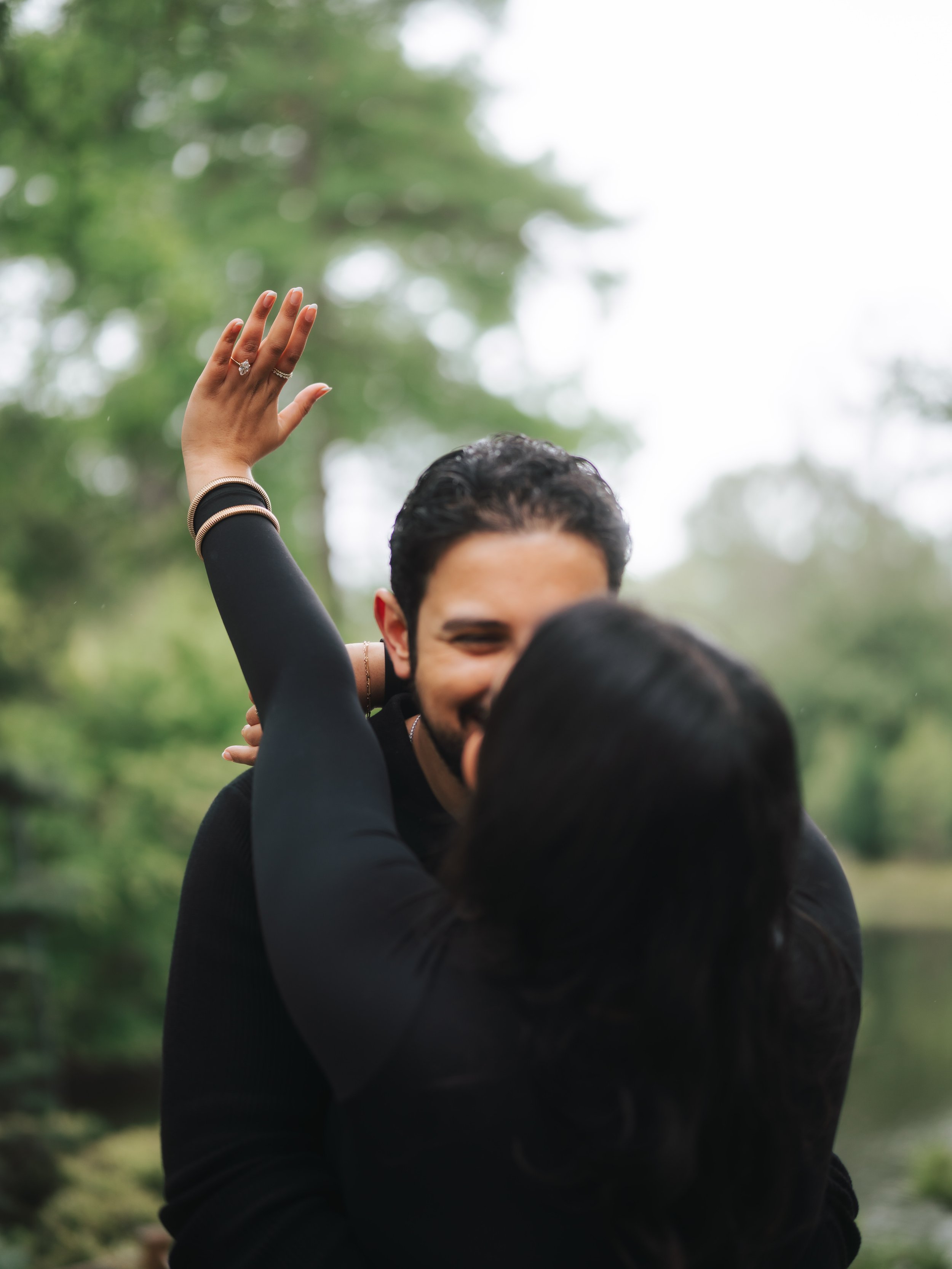  Close-up of a bride-to-be showing her marquise engagement ring with a blurred kiss in the background. 