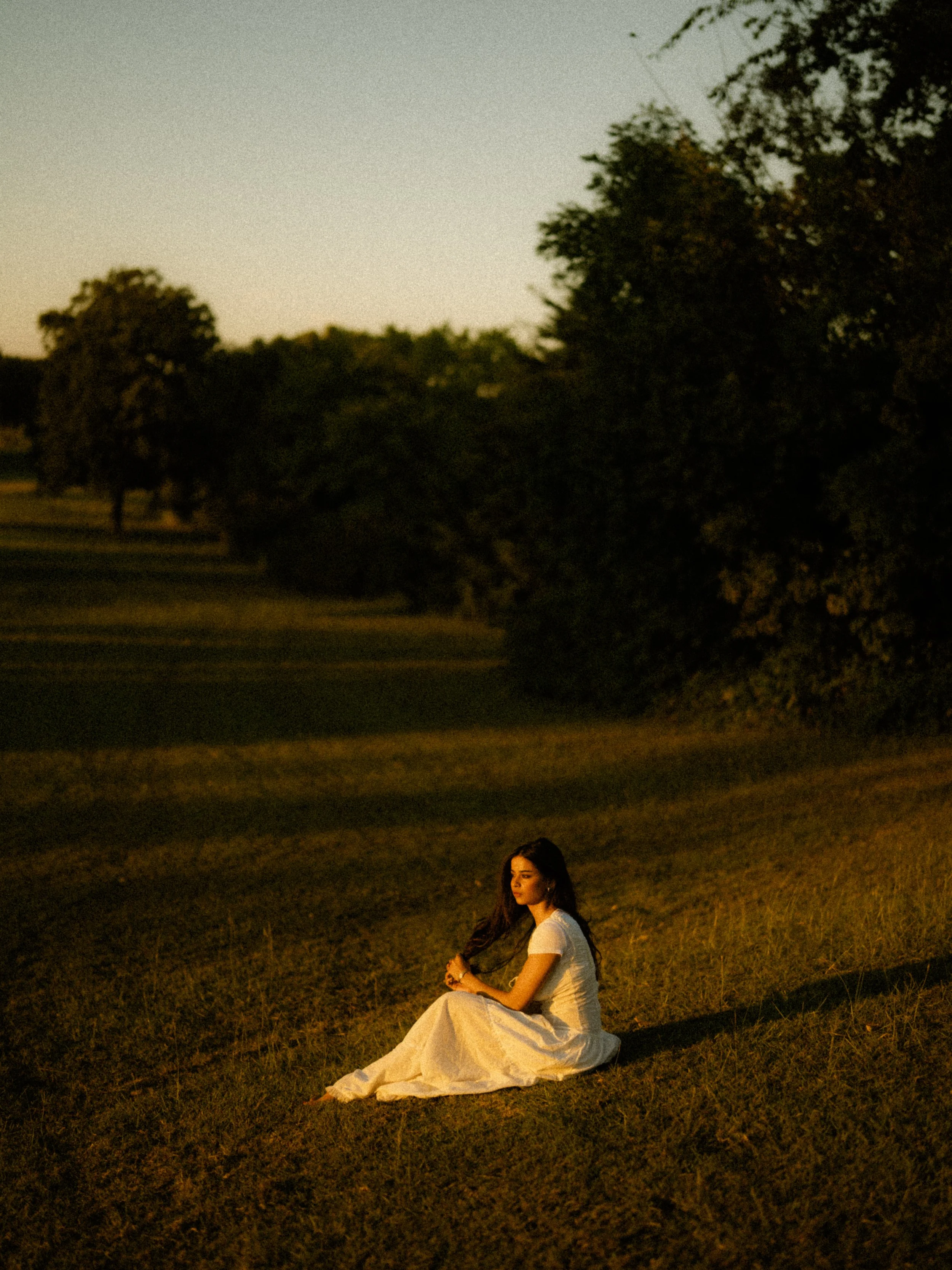  Moody film-inspired portrait of model Riya sitting in a dark field during golden hour at Breckinridge Park, Richardson, TX. Heavy grain cinematic style. 