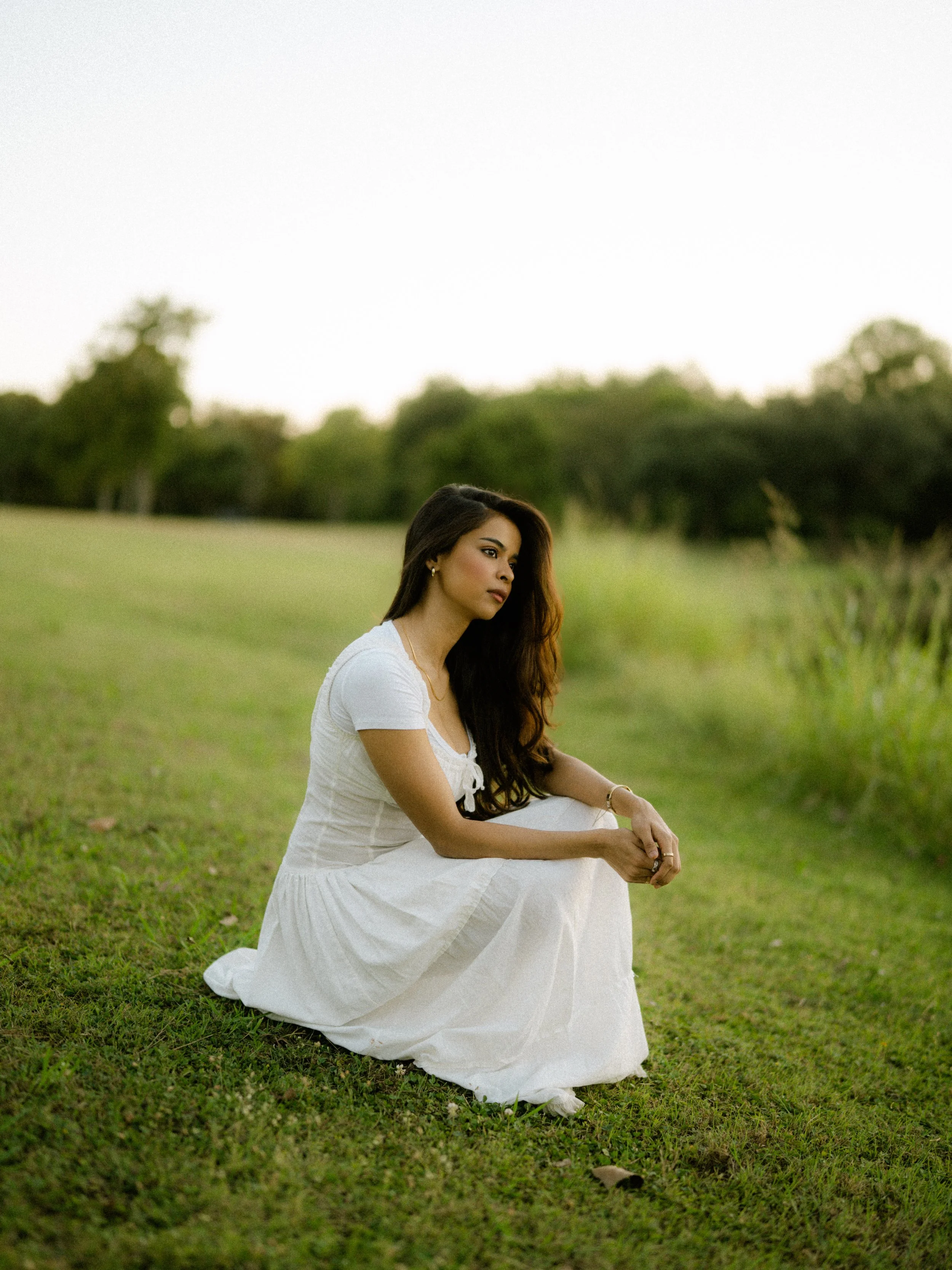  Portrait of Riya crouching in a green field at Breckinridge Park. Golden hour lighting with a natural film photography look. 