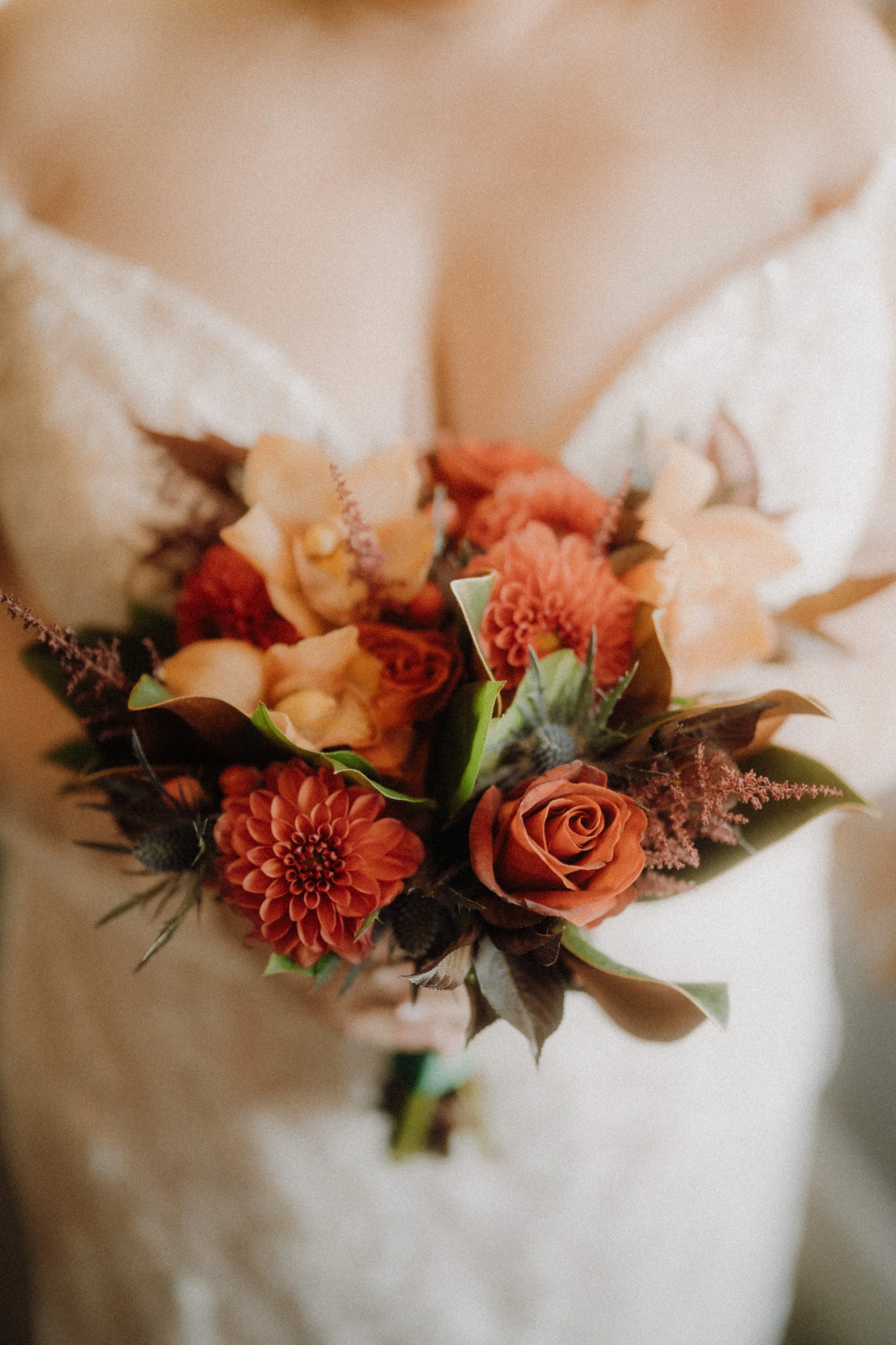 Close-up of a bride holding a bouquet of orange and red flowers, including roses, dahlias, and other blooms, with a cream dress in the background.