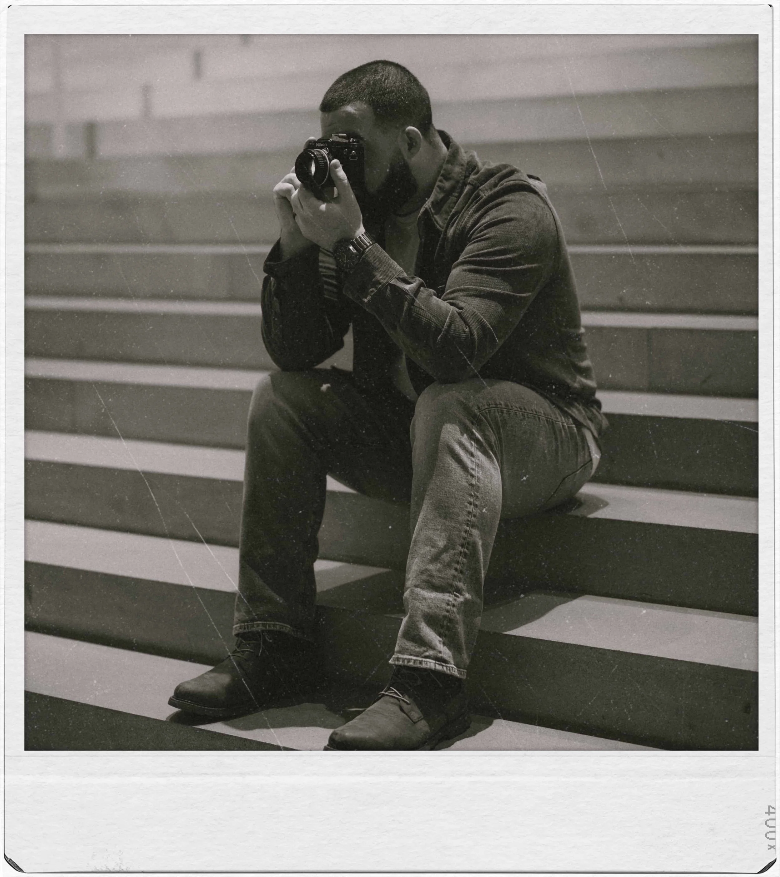 A man sitting on stairs, taking a photograph with a camera, dressed in casual dark clothing and boots, in black and white.