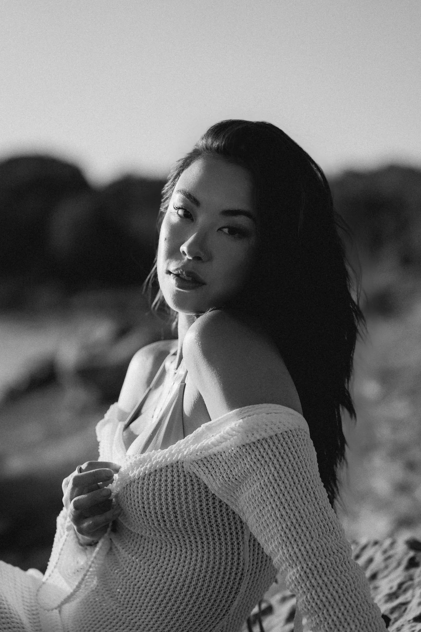 Black and white photo of a woman with long hair, looking at the camera, outdoors with rocks and blurred landscape in the background.