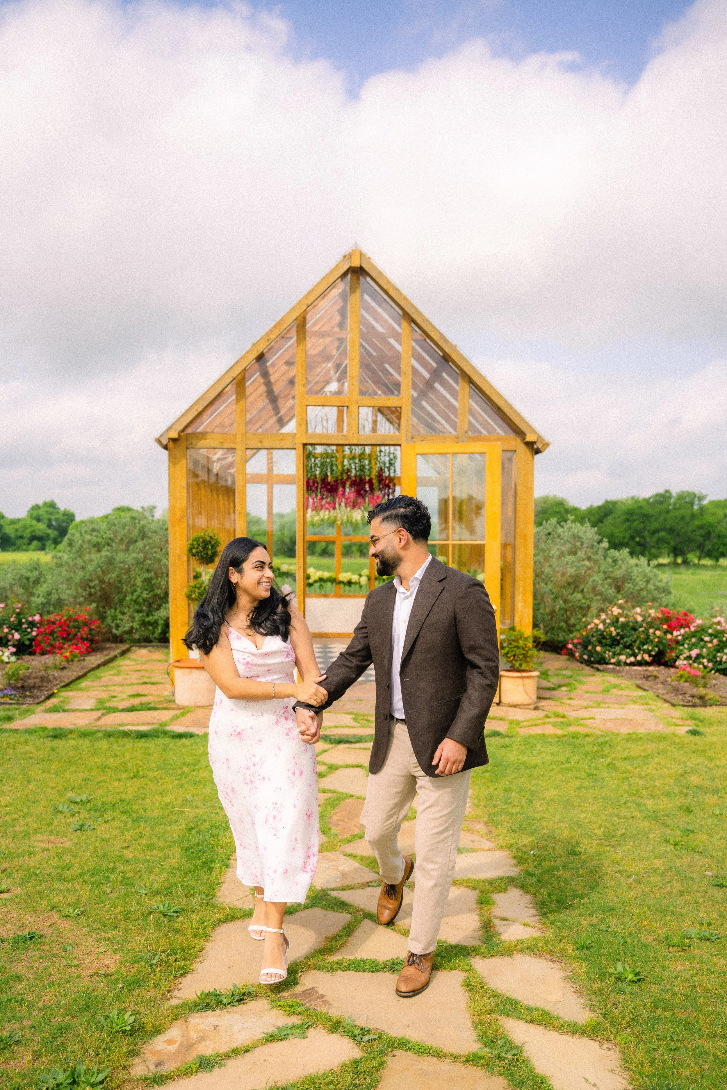 A smiling couple holding hands and walking on a stone path in front of a small glass greenhouse with flowering plants, green grass, flowers, and trees in the background.