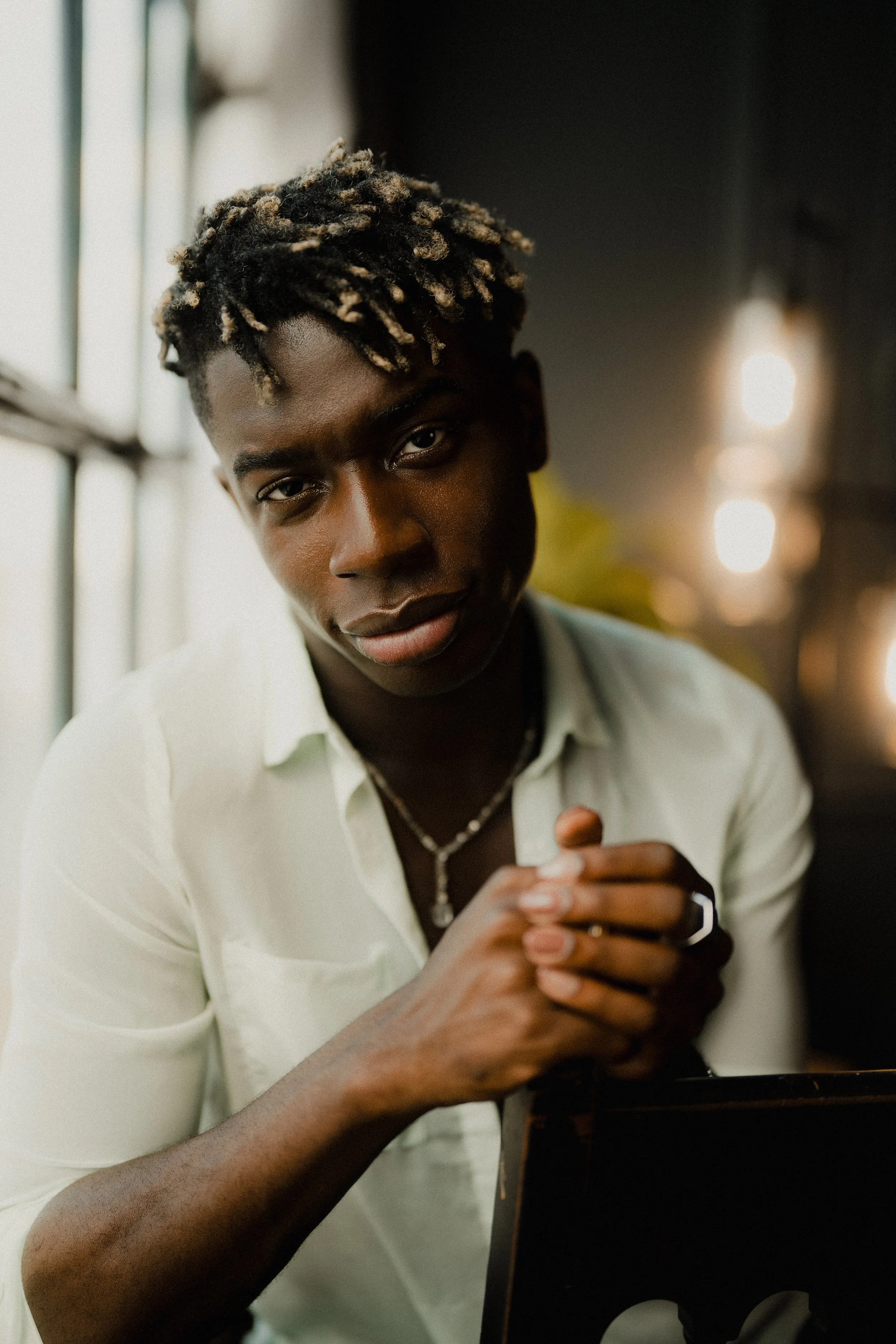 A young man with styled hair and a white shirt sitting indoors near a window, looking at the camera with hands clasped.
