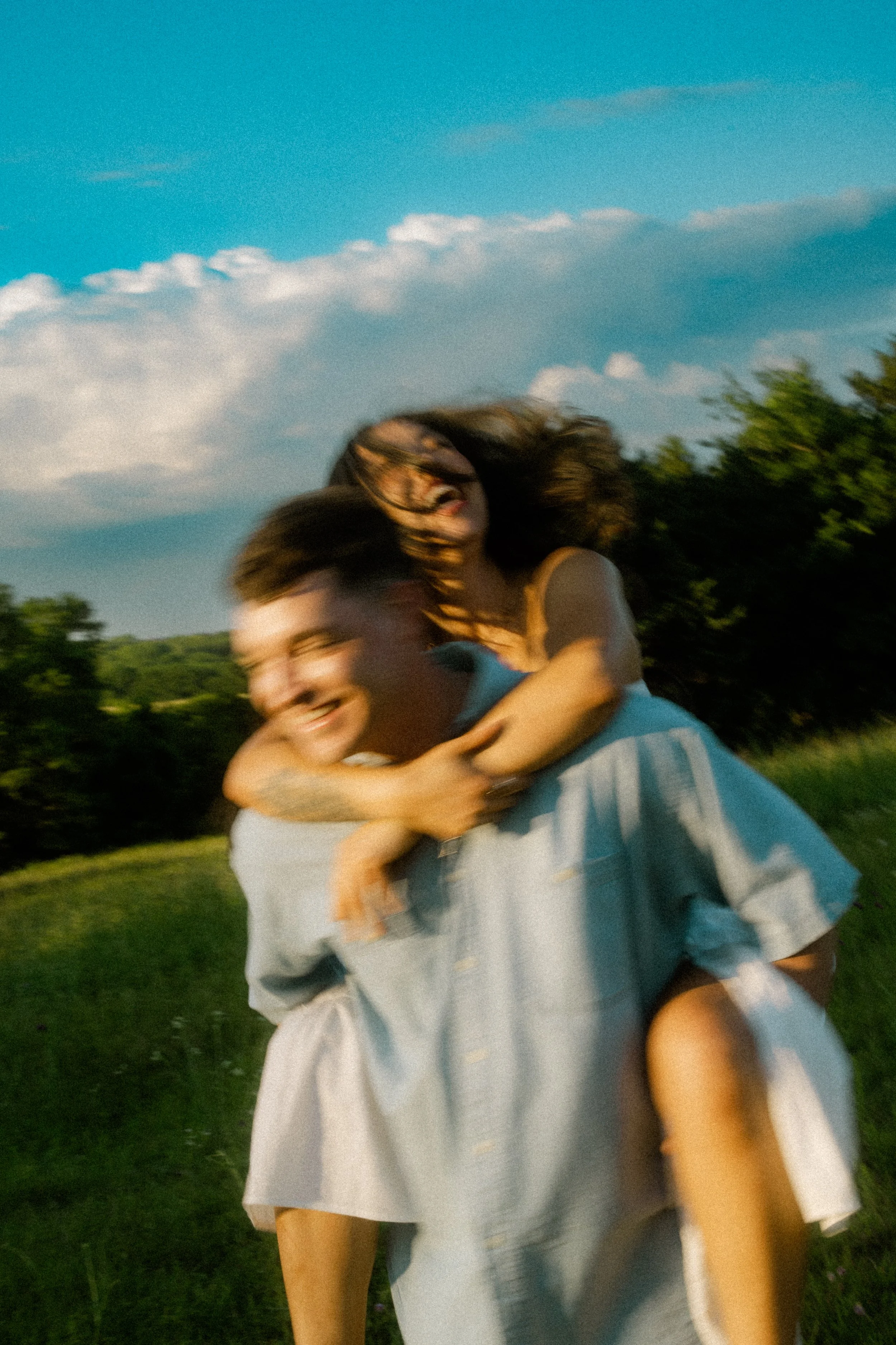 A happy couple playing outside on a sunny day, with the woman on the man's back, both smiling and laughing. They are in a grassy field with trees and blue sky in the background.