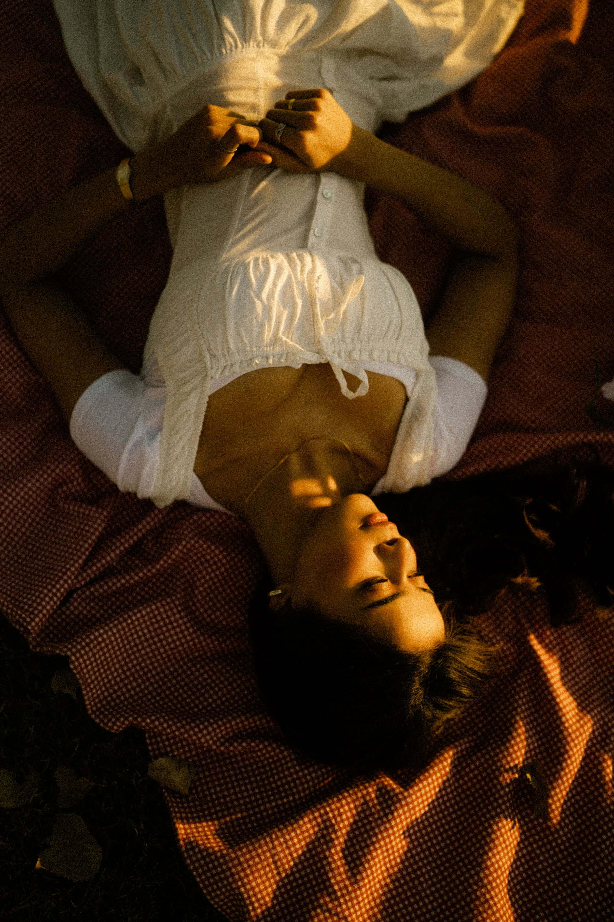 A woman lying on a bed with red and white checkered sheets, wearing a white outfit, eyes closed, enjoying the warm sunlight.