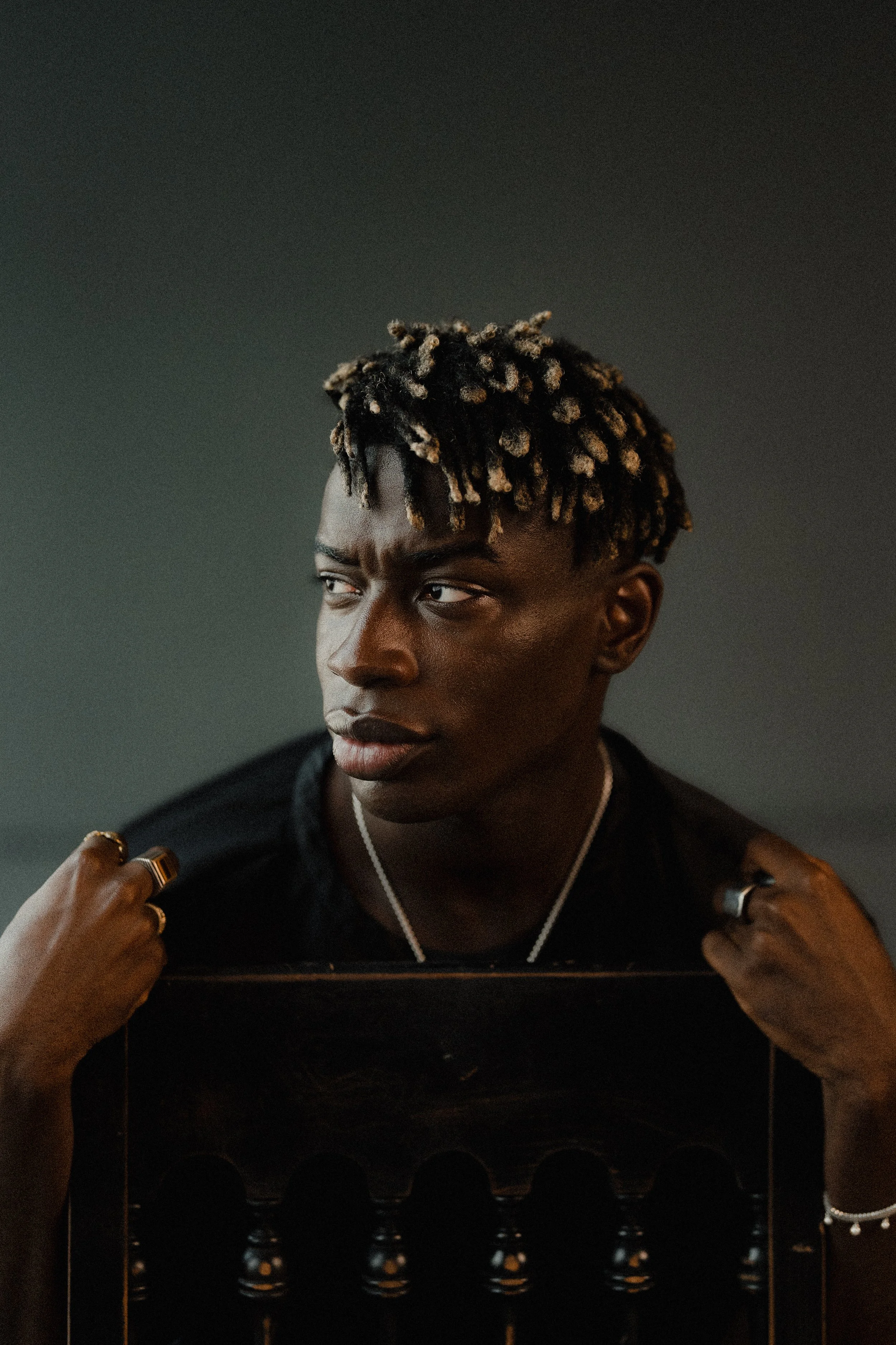 A young man with electric blonde dreadlocks is looking off to the side with a serious expression, holding a dark piece of furniture with both hands, against a plain, dark background.