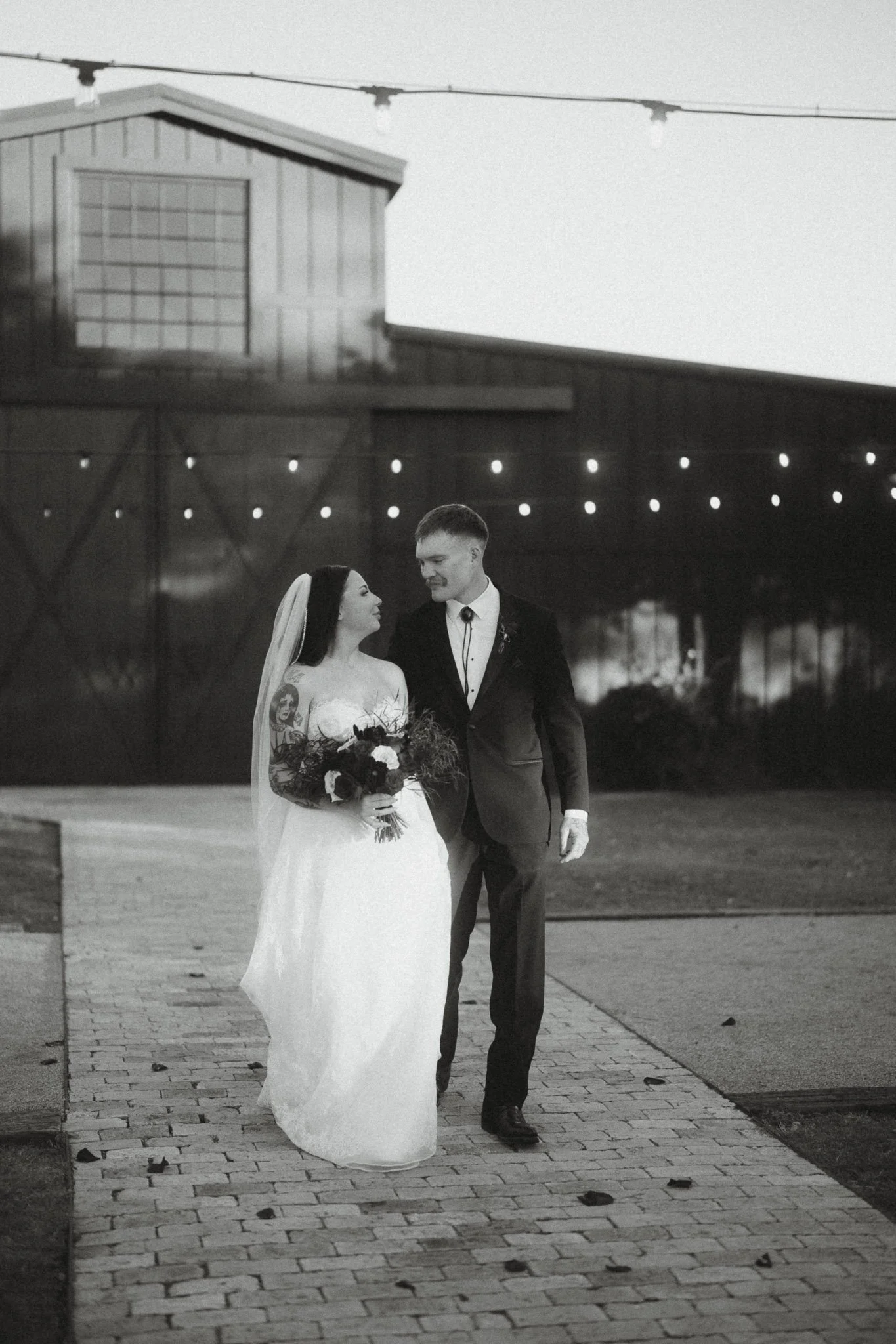 Black and white photo of a bride and groom walking together outdoors, with string lights overhead, a barn-like building in the background, and the bride holding a bouquet of flowers.