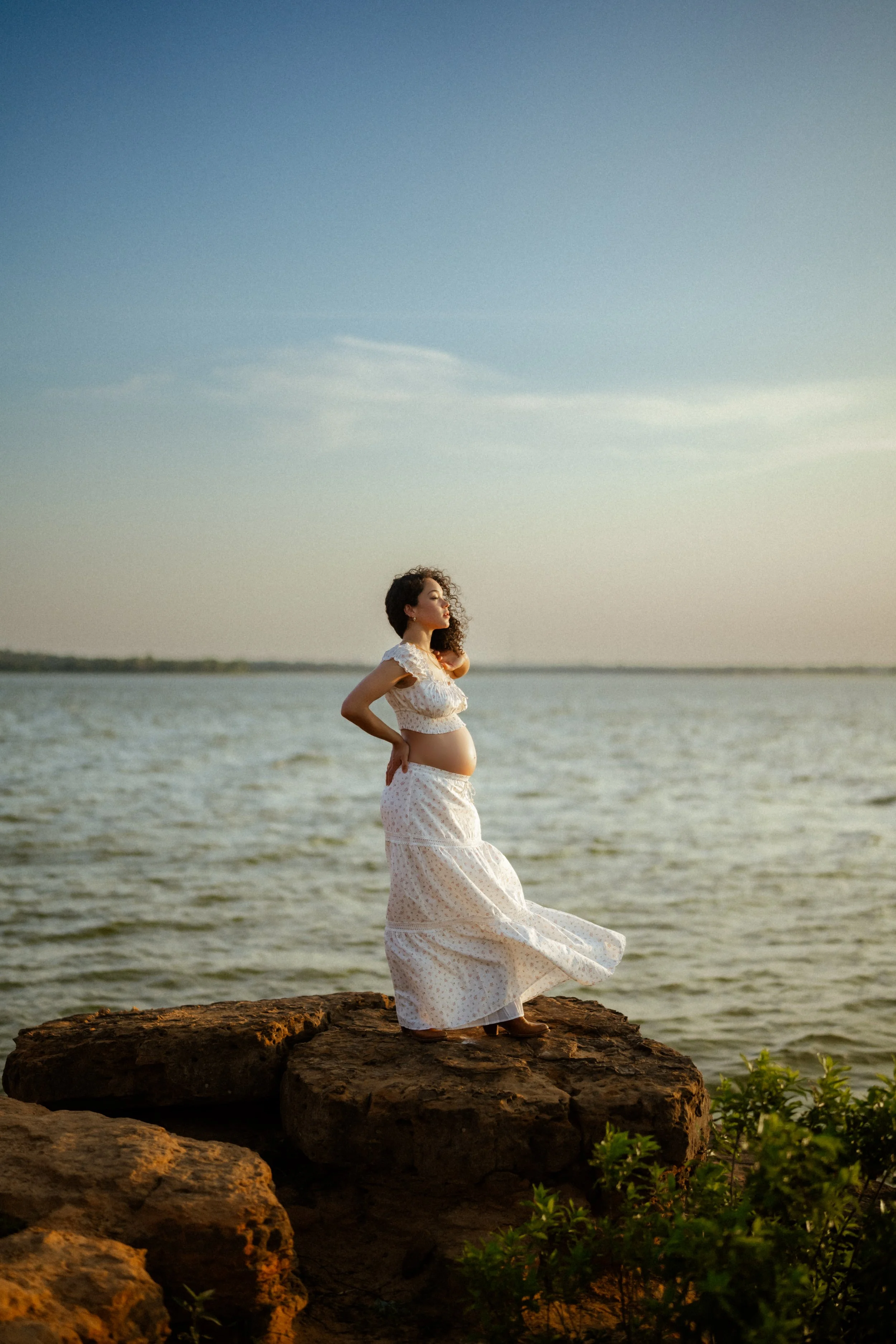 Pregnant woman in a white dress standing on rocks by the water at sunset.