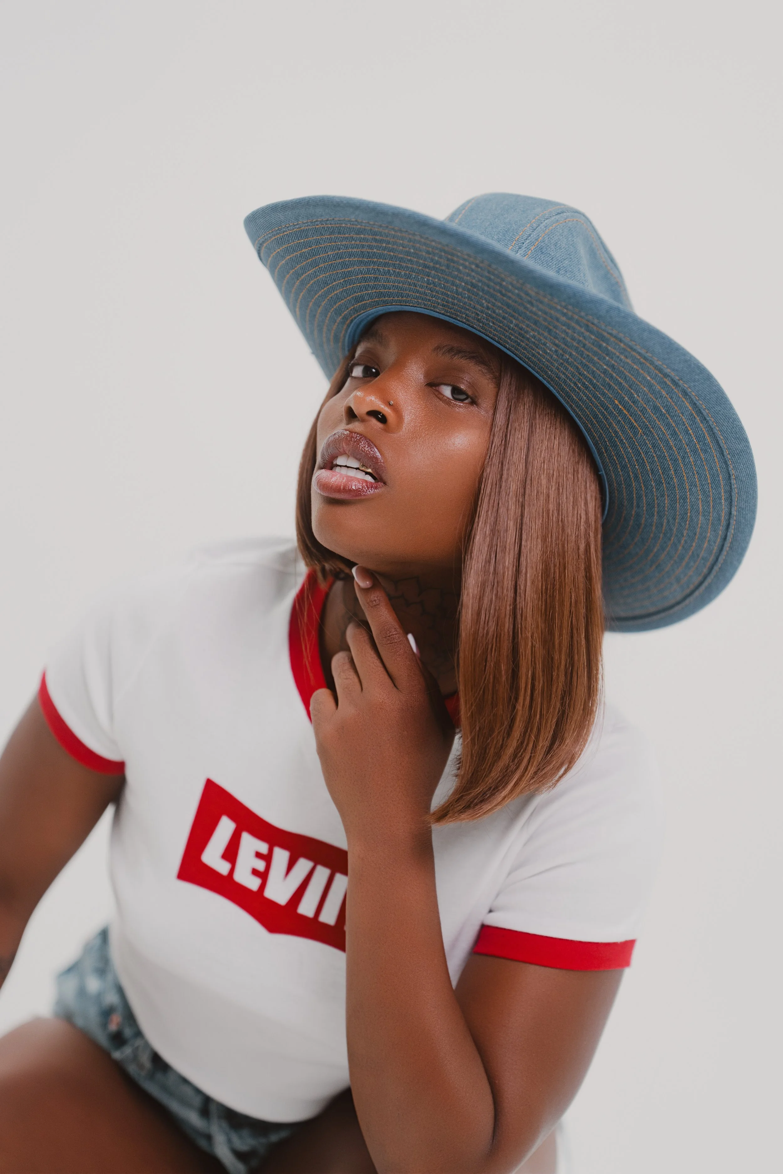 A woman wearing a large blue hat, a white Levi's t-shirt with red trim, and denim shorts, posing against a plain white background.