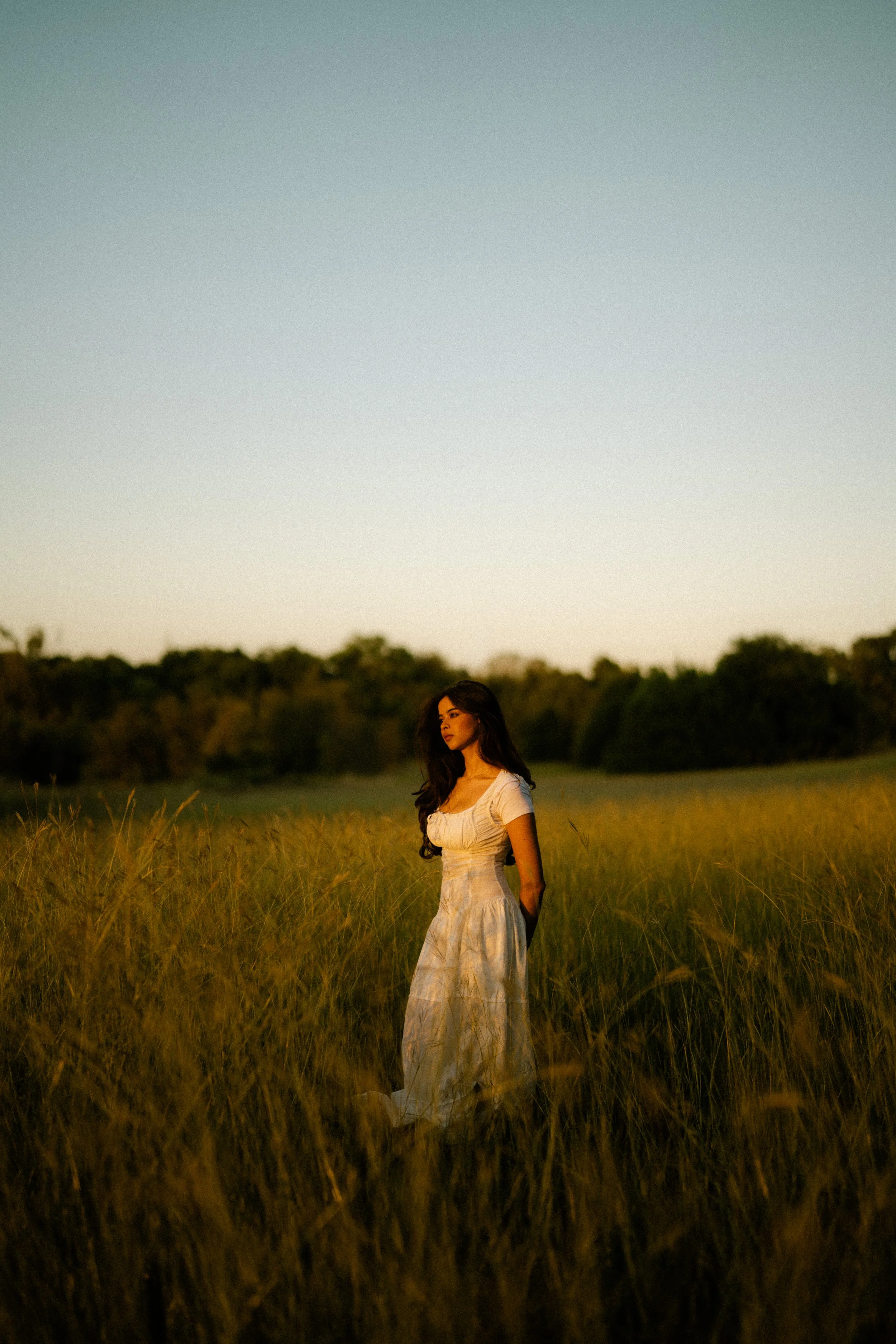 A woman in a white dress standing in a field of tall grass during sunset.
