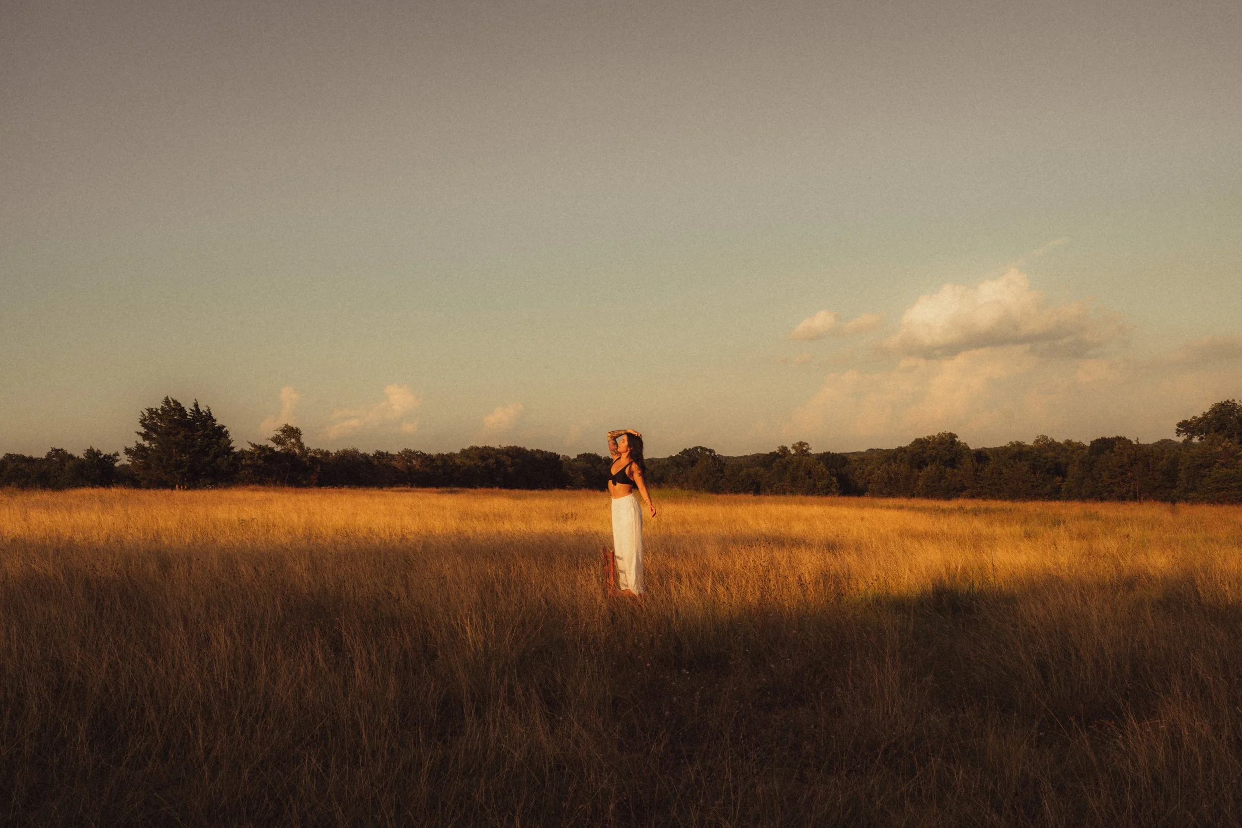 Woman standing in a grassy field during sunset, looking into the distance with one hand on her forehead and wearing a black crop top and white pants.