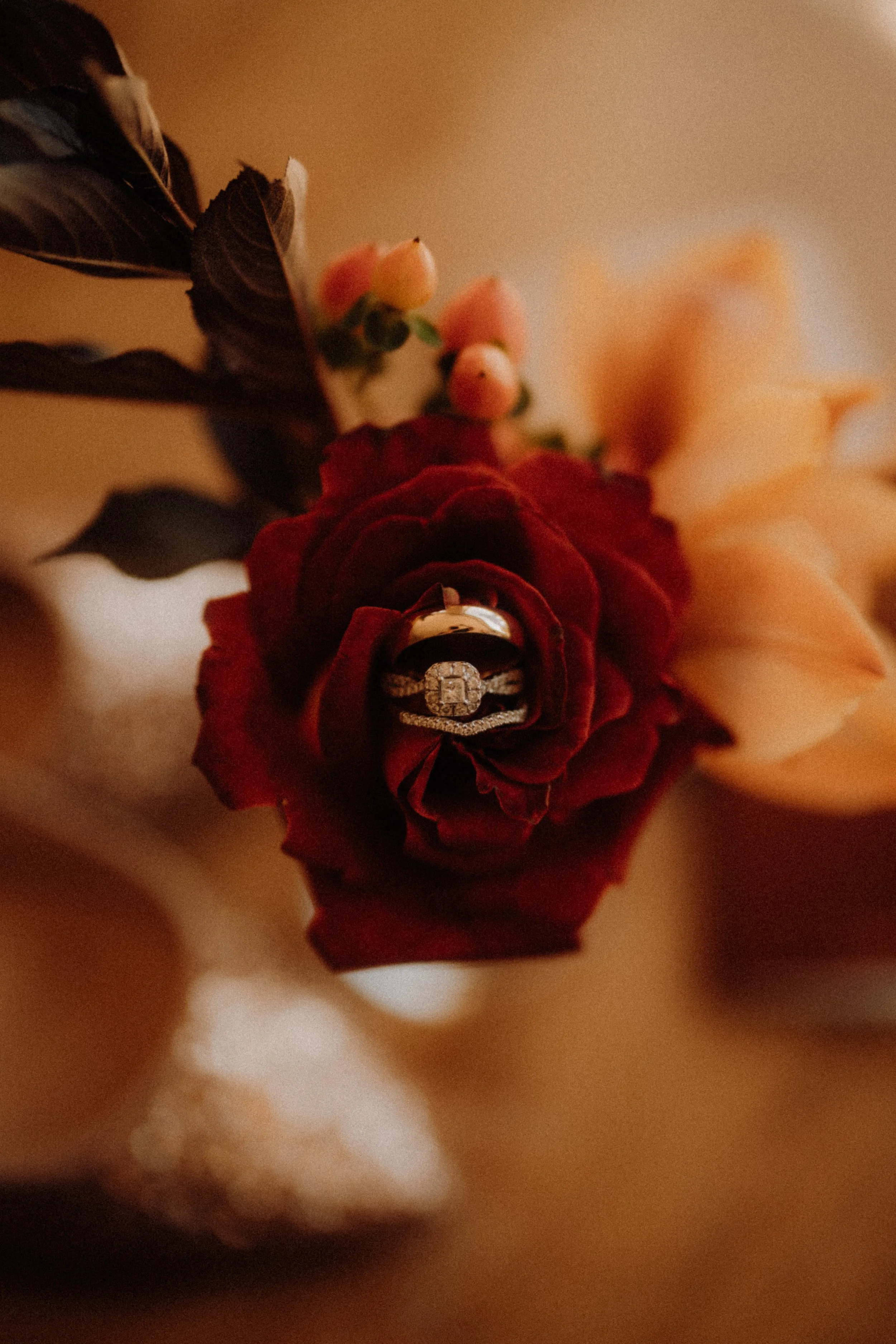 Close-up of a bouquet with a red flower that has a diamond engagement ring and wedding band placed inside its petals. The bouquet includes small pink and peach-colored flowers and dark green leaves, with a warm, blurred background.