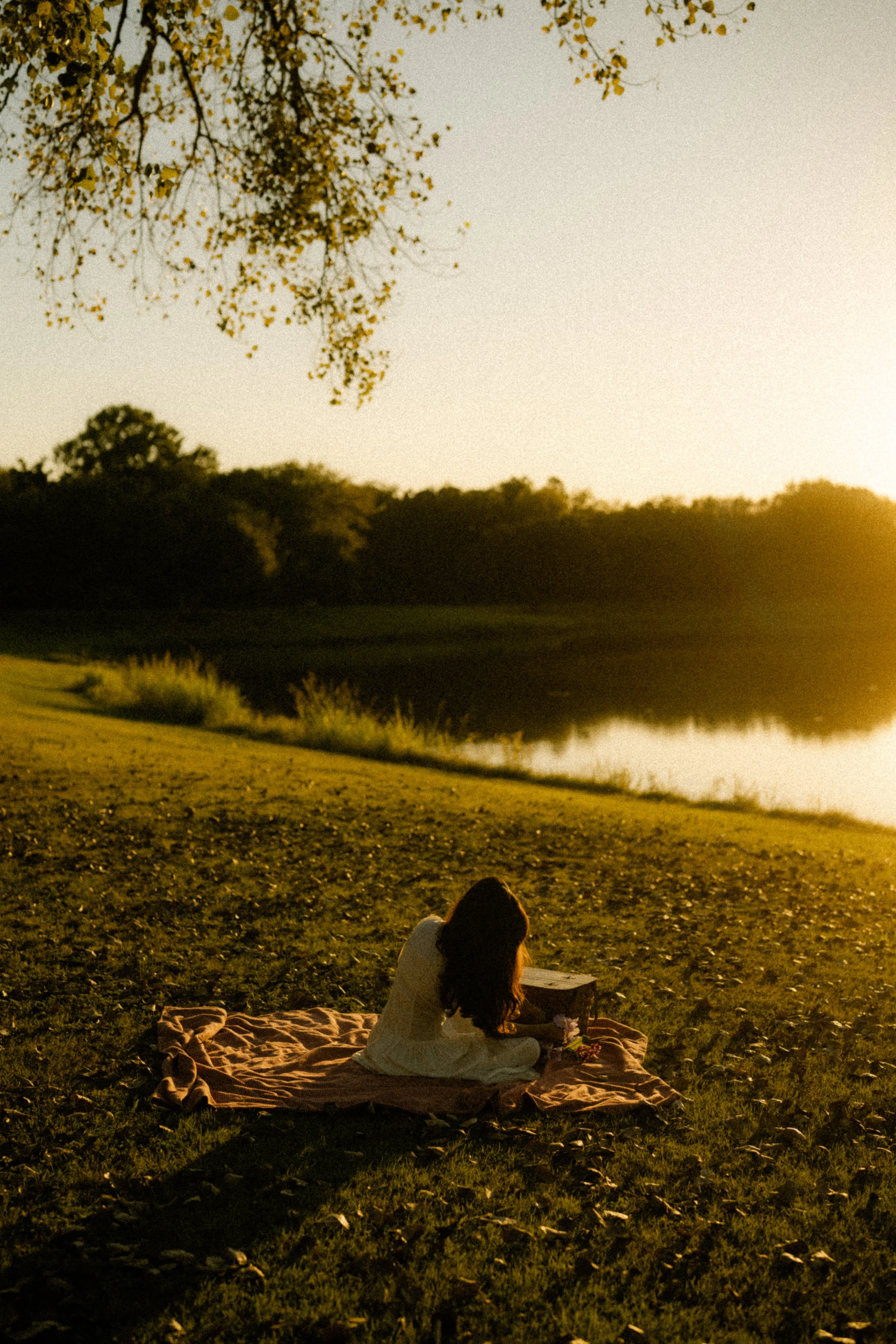 A woman with long hair sitting on a blanket near a lake during sunset, reading a book.