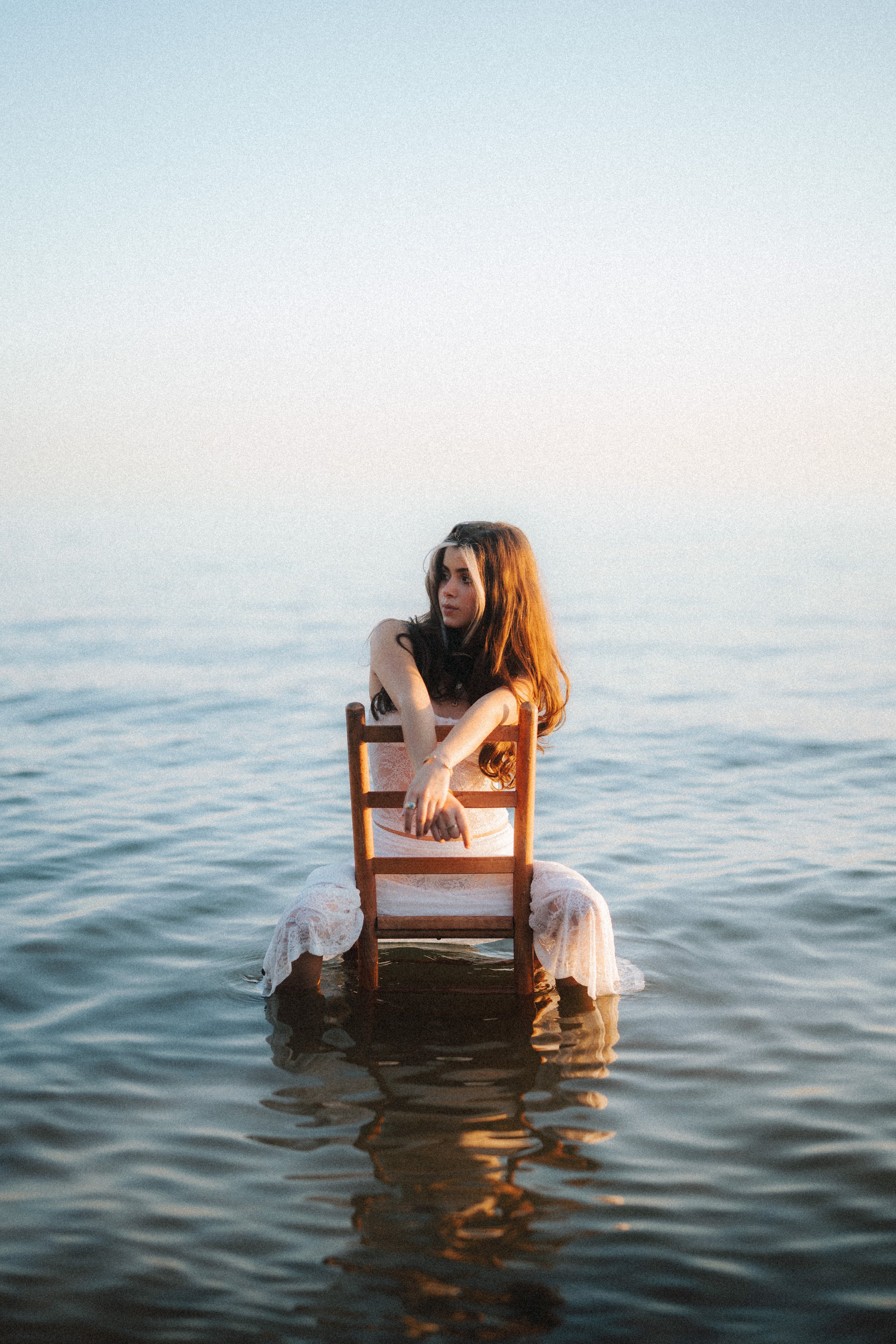 A woman with long brown hair wearing a white dress is sitting on a wooden chair in the water, facing slightly left with her arms resting on the chair's back.