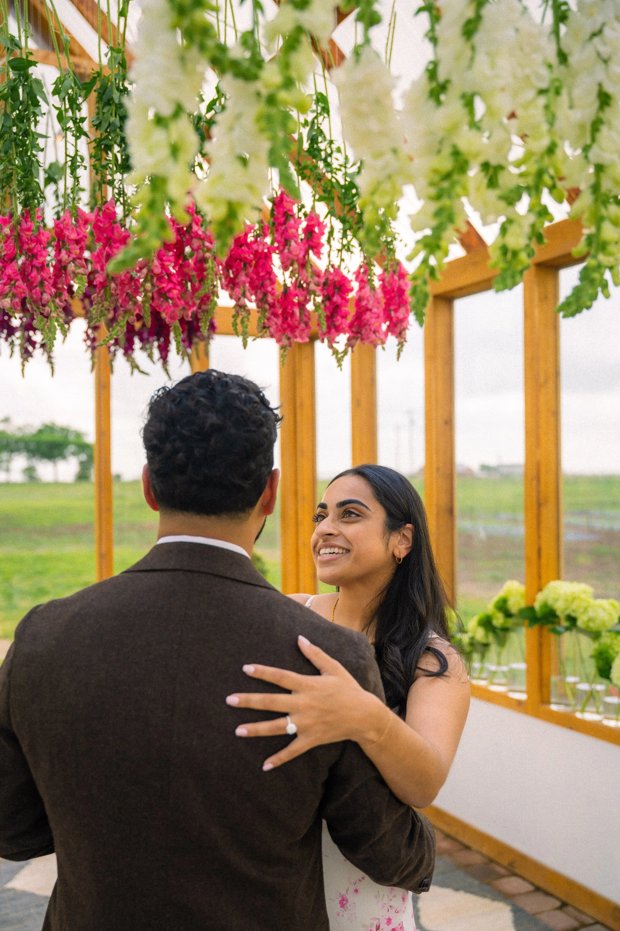 A woman and man are dancing and smiling in an outdoor setting, decorated with hanging pink and white flowers and greenery.