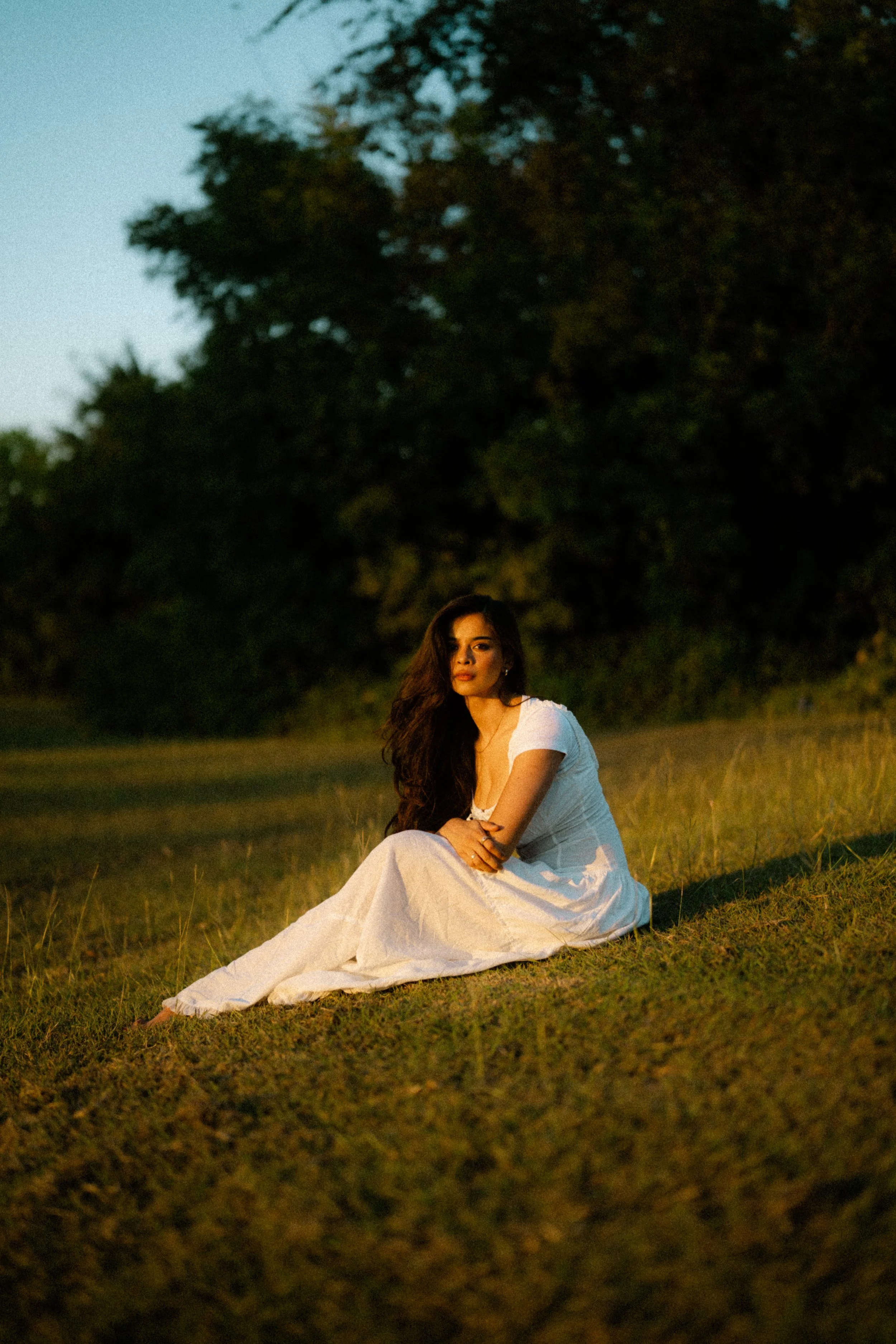A woman in a white dress sitting on grass during sunset with trees in the background.