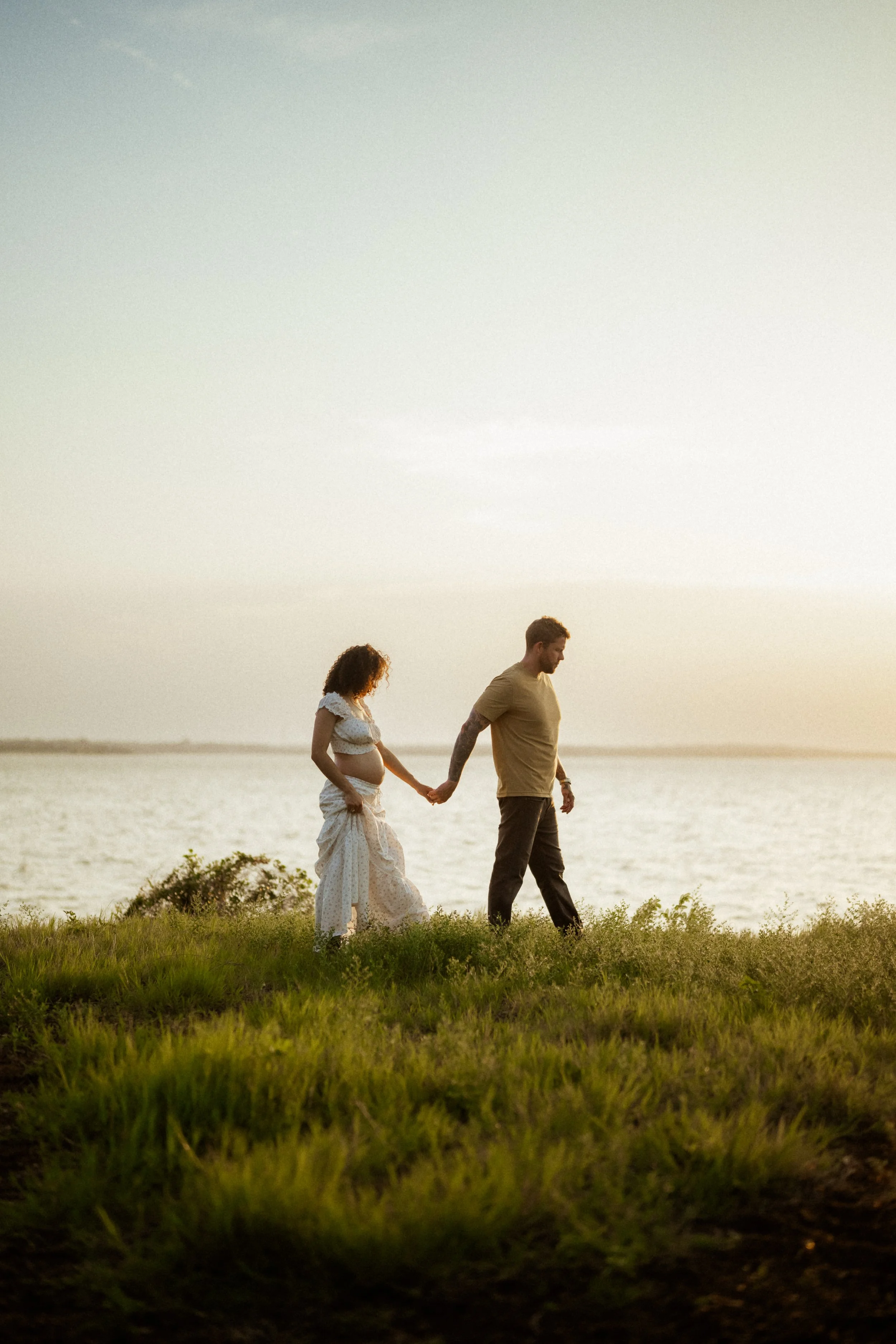 A pregnant woman and a man holding hands walking on grass near a body of water at sunset.