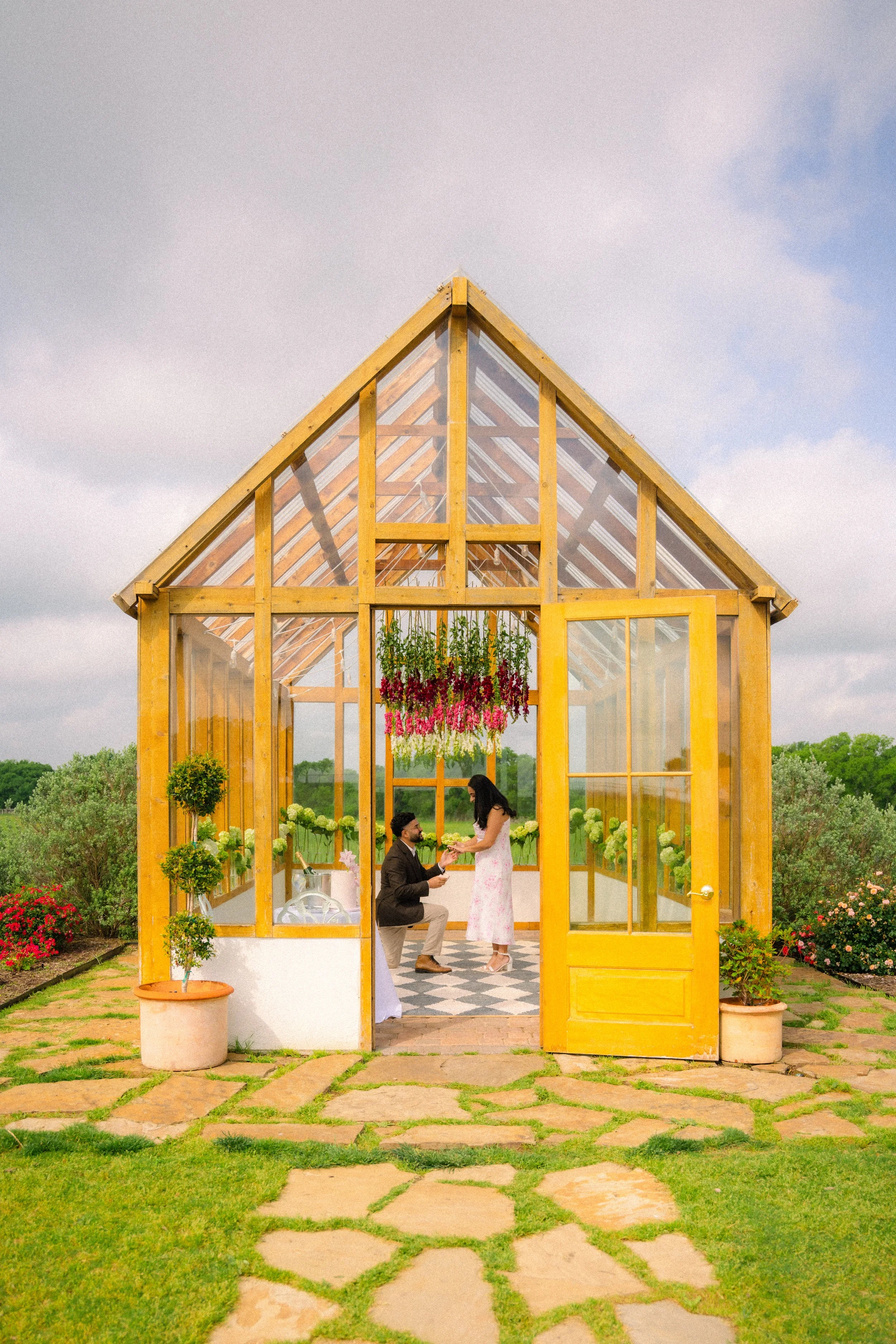 A man proposing to a woman inside a glass greenhouse with potted plants and hanging flowers, surrounded by a garden.