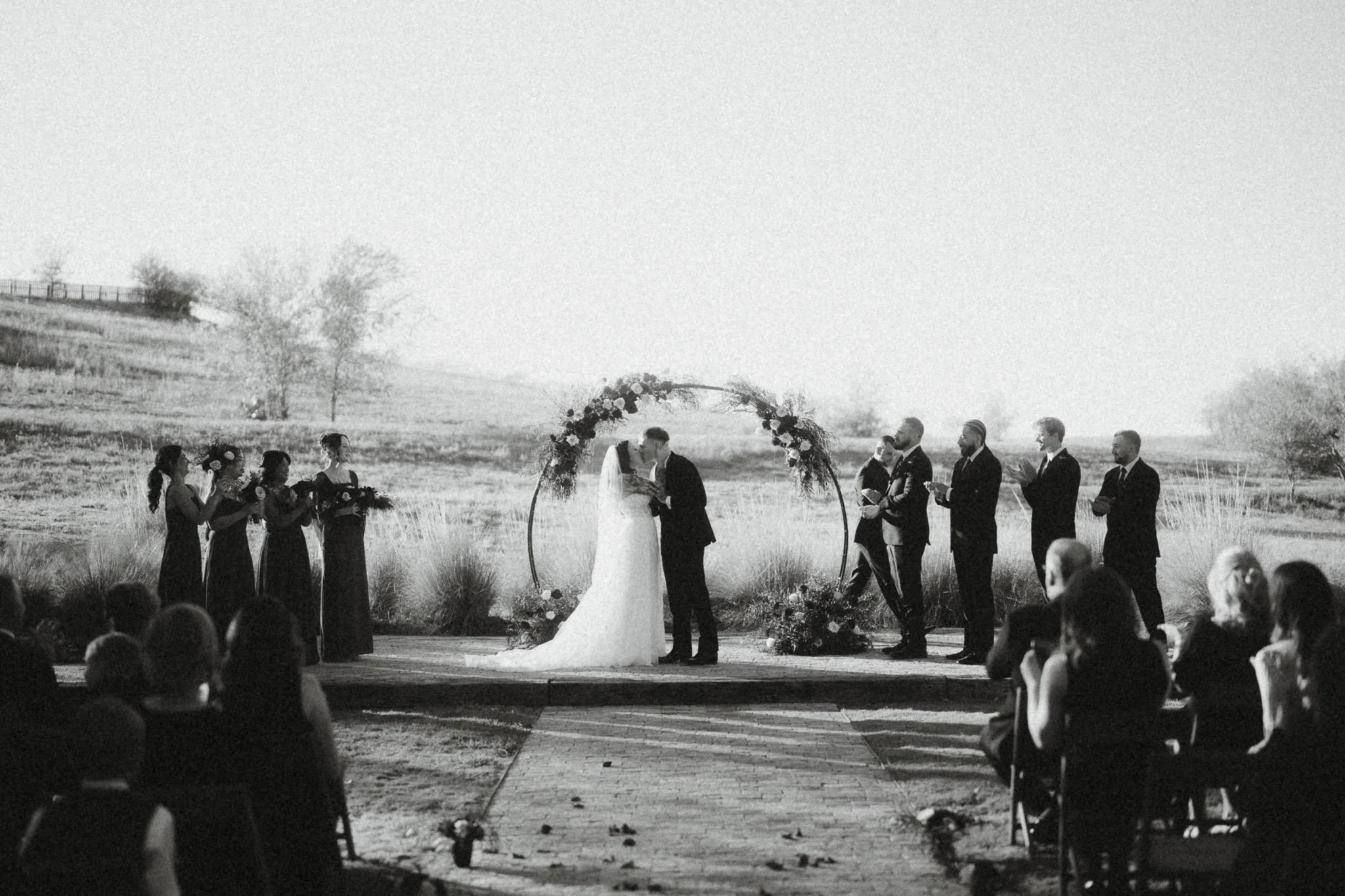 Black and white photograph of a wedding ceremony outdoors, featuring a bride and groom kissing under a floral arch with the officiant, bridesmaids, and groomsmen nearby, and guests seated watching.