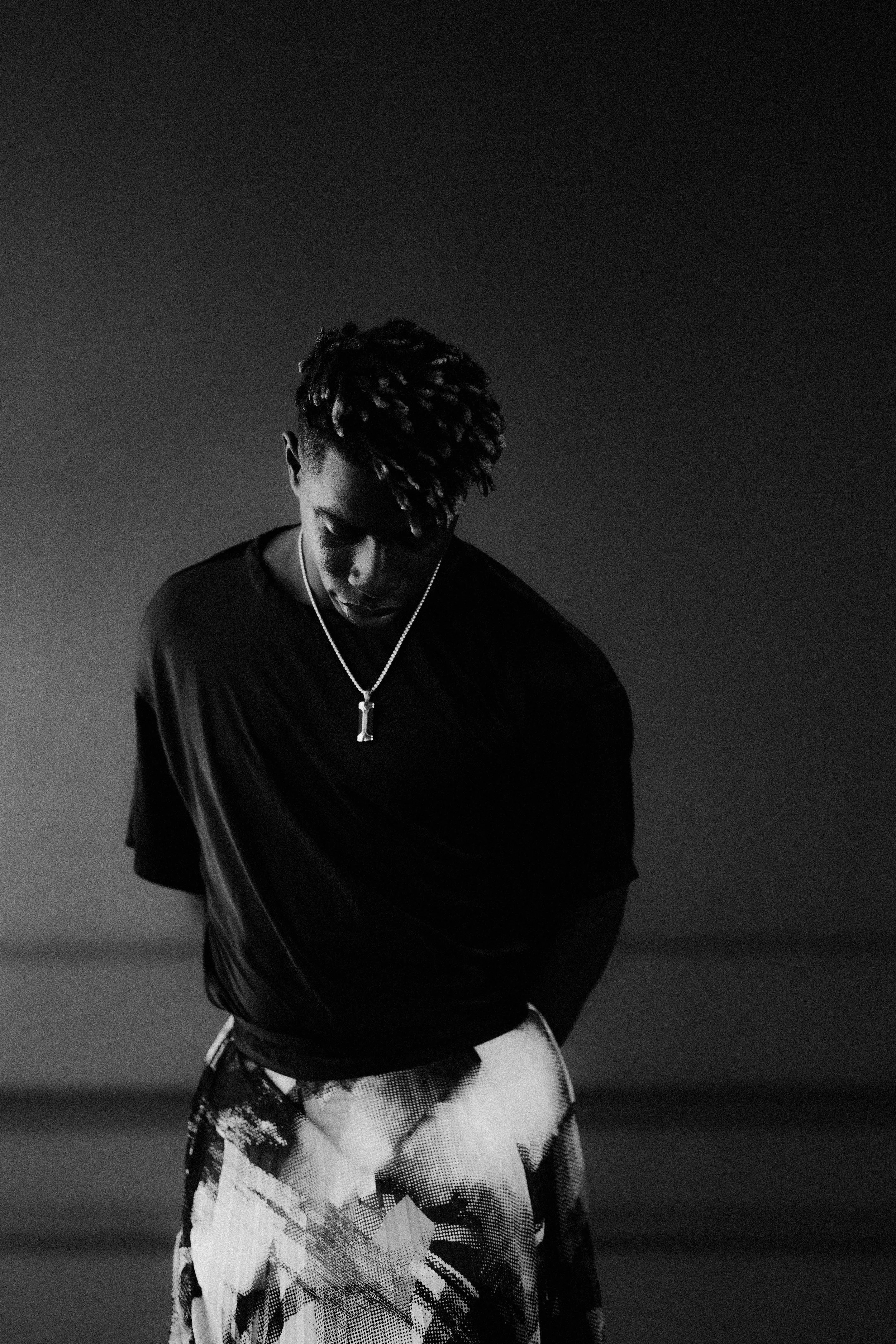 A black and white photo of a young man with dreadlocks, wearing a black t-shirt and patterned pants, looking down with his hands behind his back.