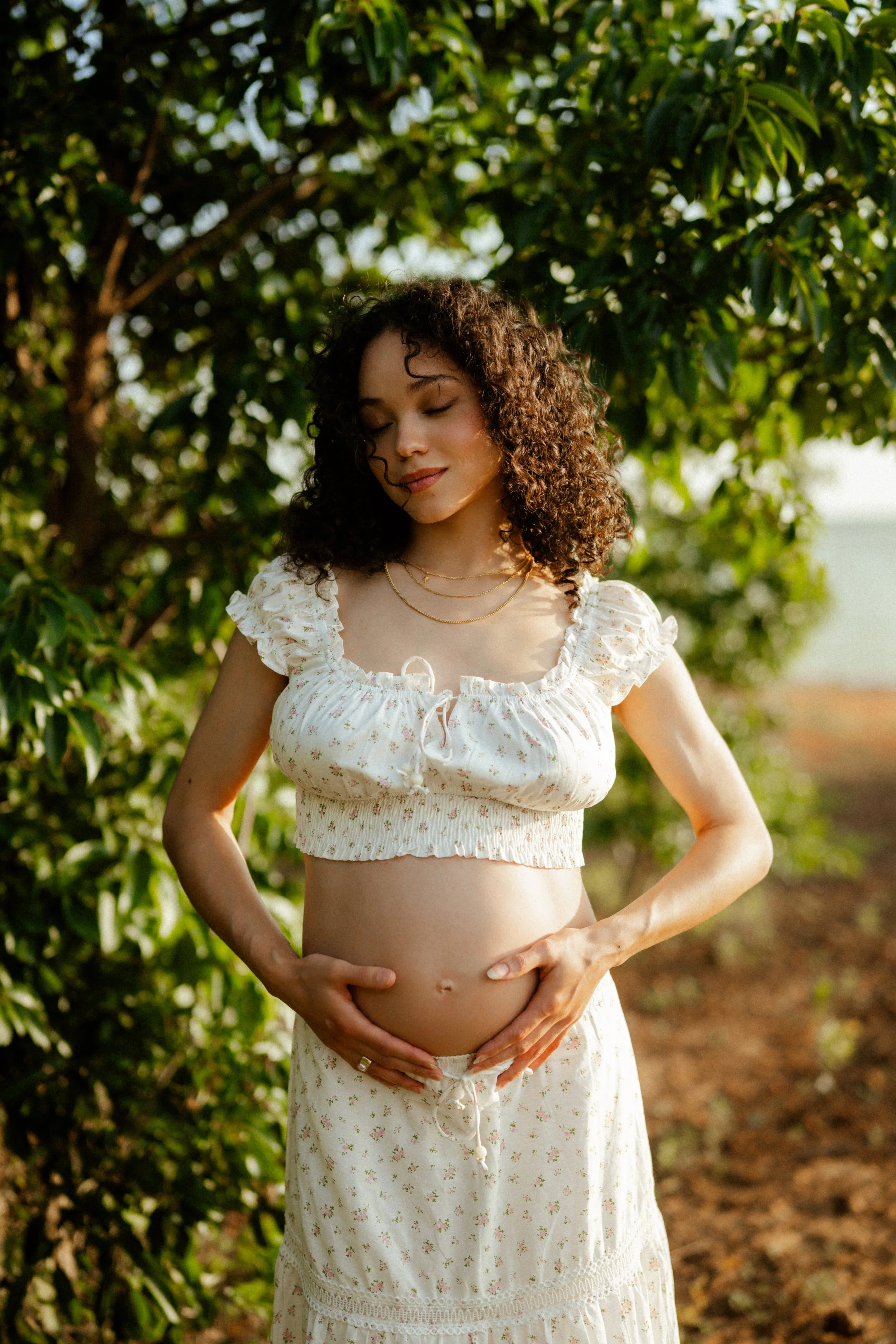 A pregnant woman with curly brown hair, wearing a white floral crop top and matching skirt, standing outdoors surrounded by green trees and foliage, gently holding her belly with both hands and smiling softly.