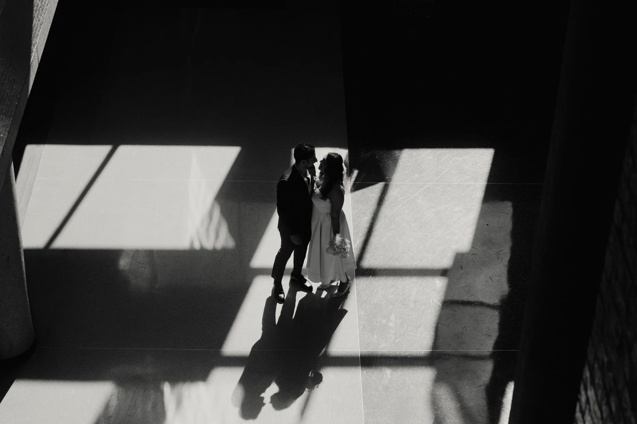 A couple in wedding attire standing close together, holding hands, with their reflections visible on the shiny floor, illuminated by sunlight through large window panes casting geometric shadows in a modern indoor setting.