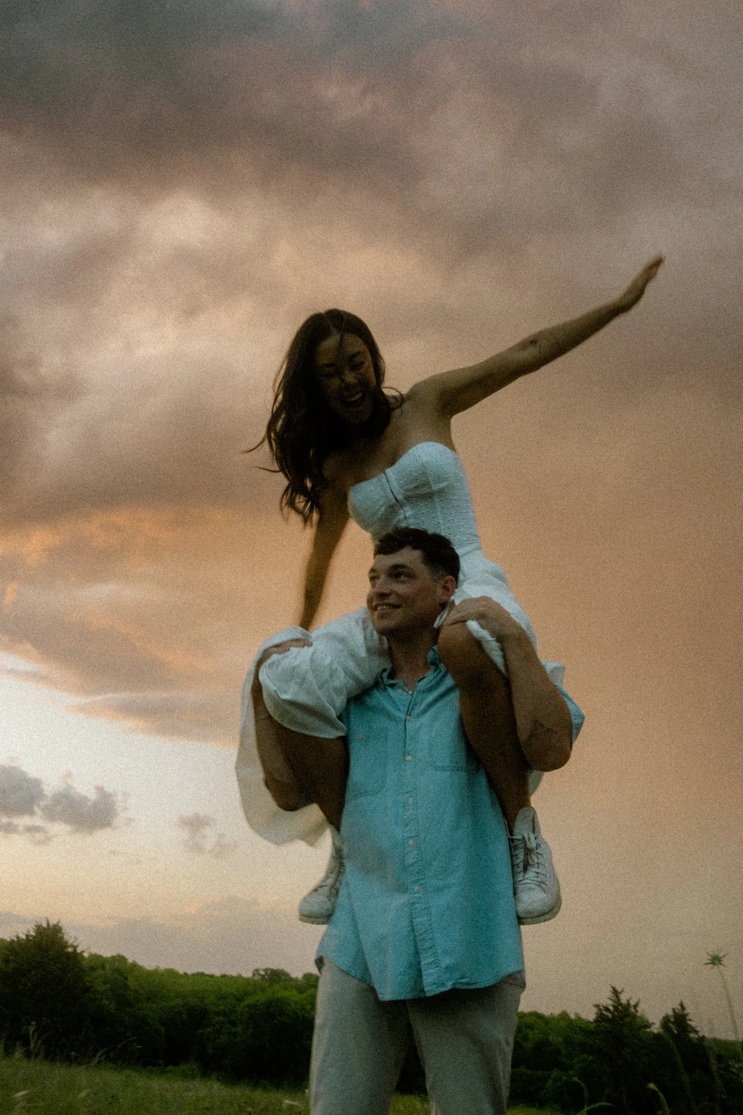 A couple outdoors at sunset, the woman sitting on the man's shoulders, both smiling, with a scenic grassy landscape and cloudy sky in the background.