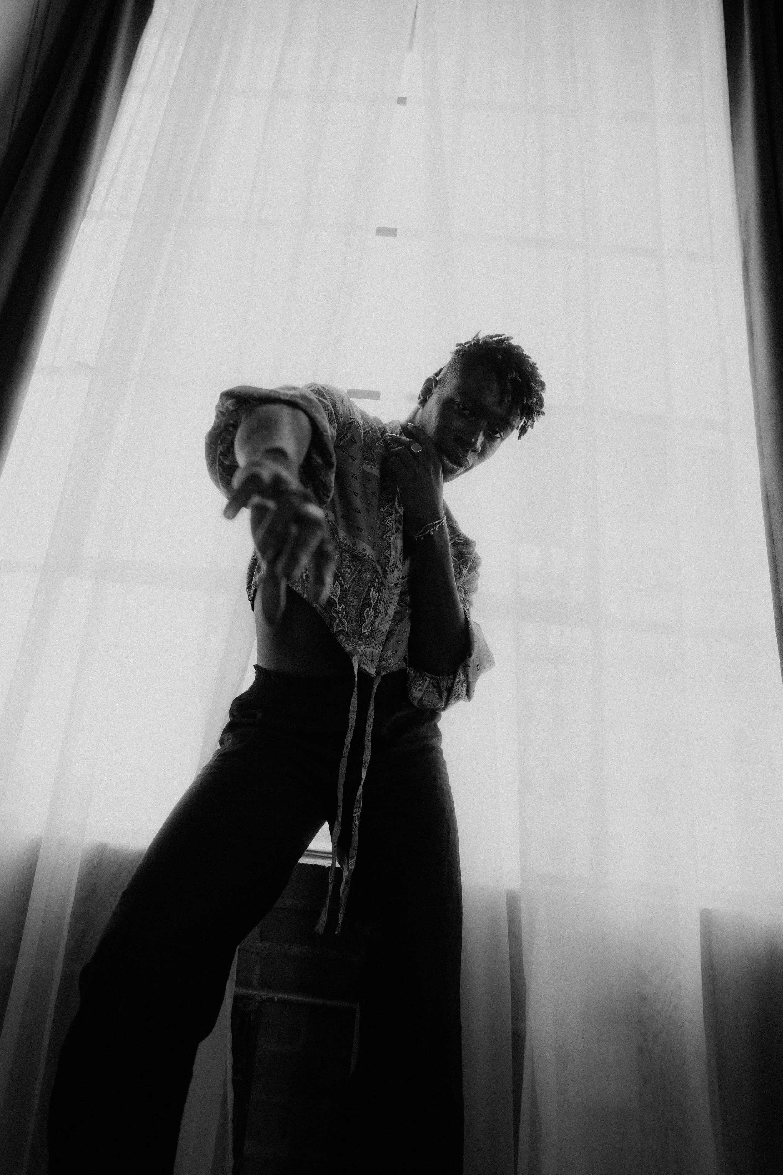 A black and white photo of a person standing in front of a sheer curtain, looking down at the camera, with one hand near their face and the other extended toward the camera.
