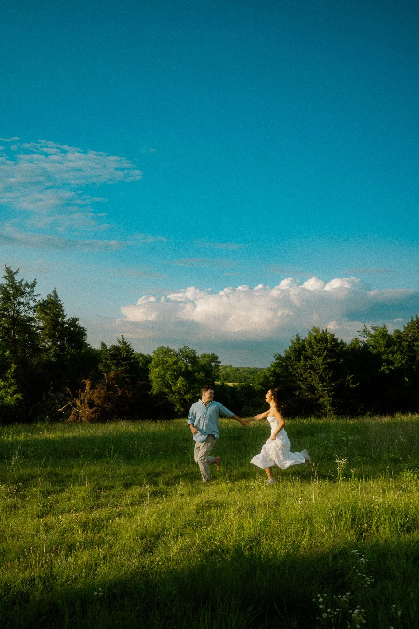Dallas, Texas couples session with a couple dancing and holding hands in a grassy field outdoors under a clear blue sky with clouds, trees in the background, during daylight.