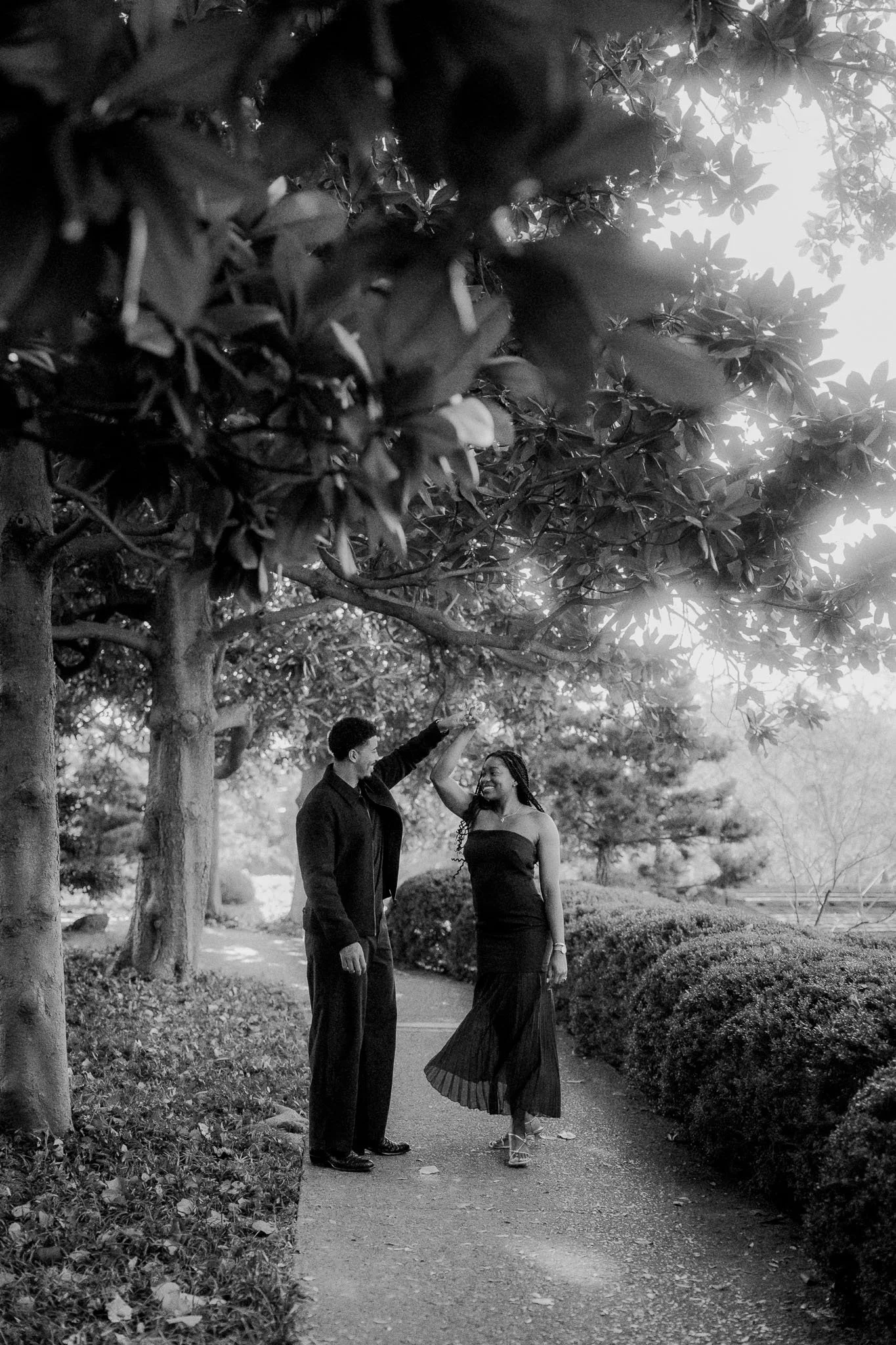 Couple dancing or celebrating on a park pathway surrounded by trees and bushes, smiling and enjoying each other's company in black and white.