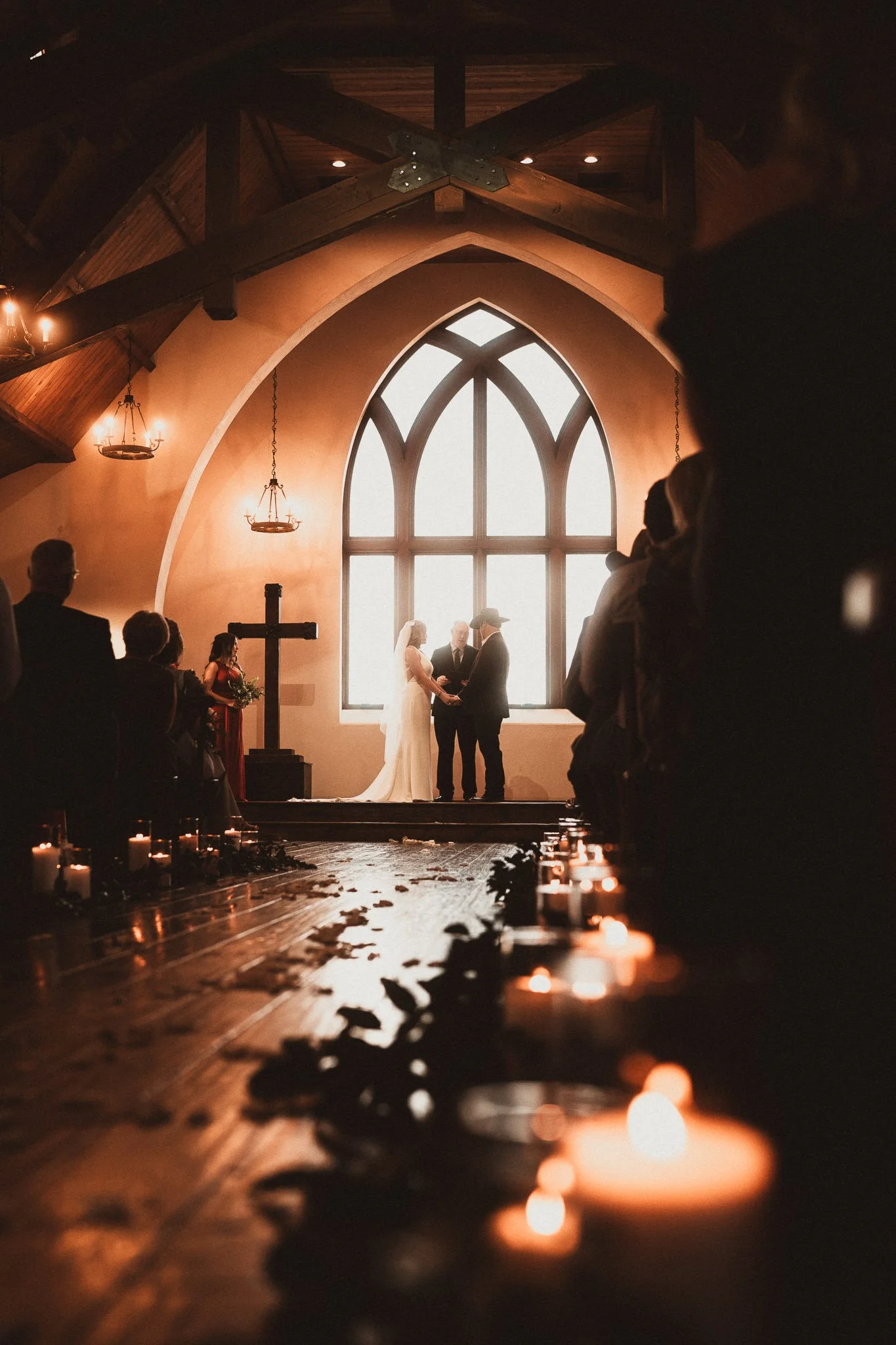 A wedding ceremony taking place inside a church with a large arched window as the backdrop. The bride and groom are holding hands with officiant in between, facing each other at the altar. Guests are seated along the aisle, illuminated by lit candles on the floor.