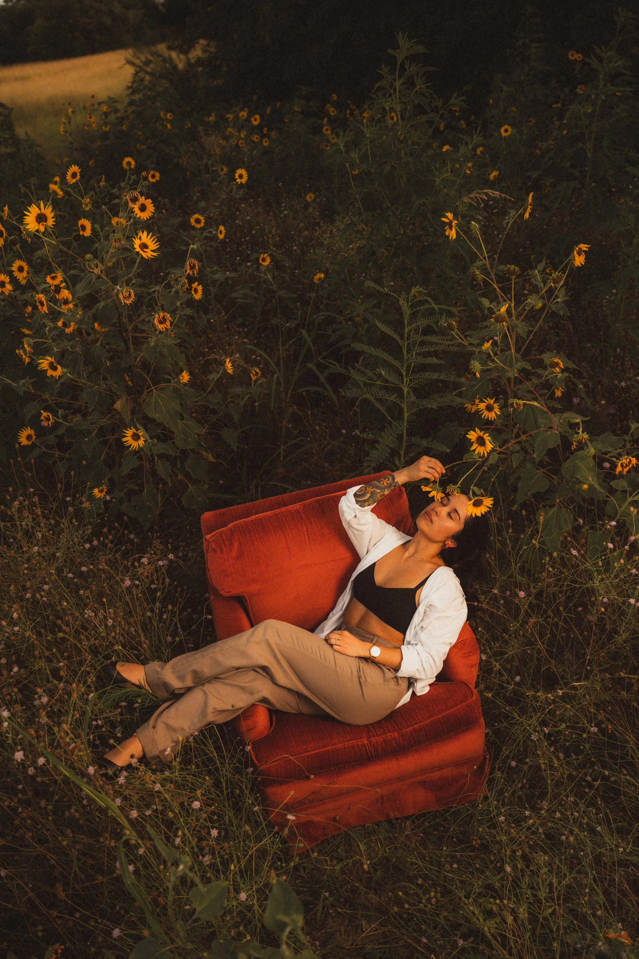 A woman relaxing on a red velvet armchair in a field of sunflowers at dusk, with her eyes closed and holding a sunflower near her face.